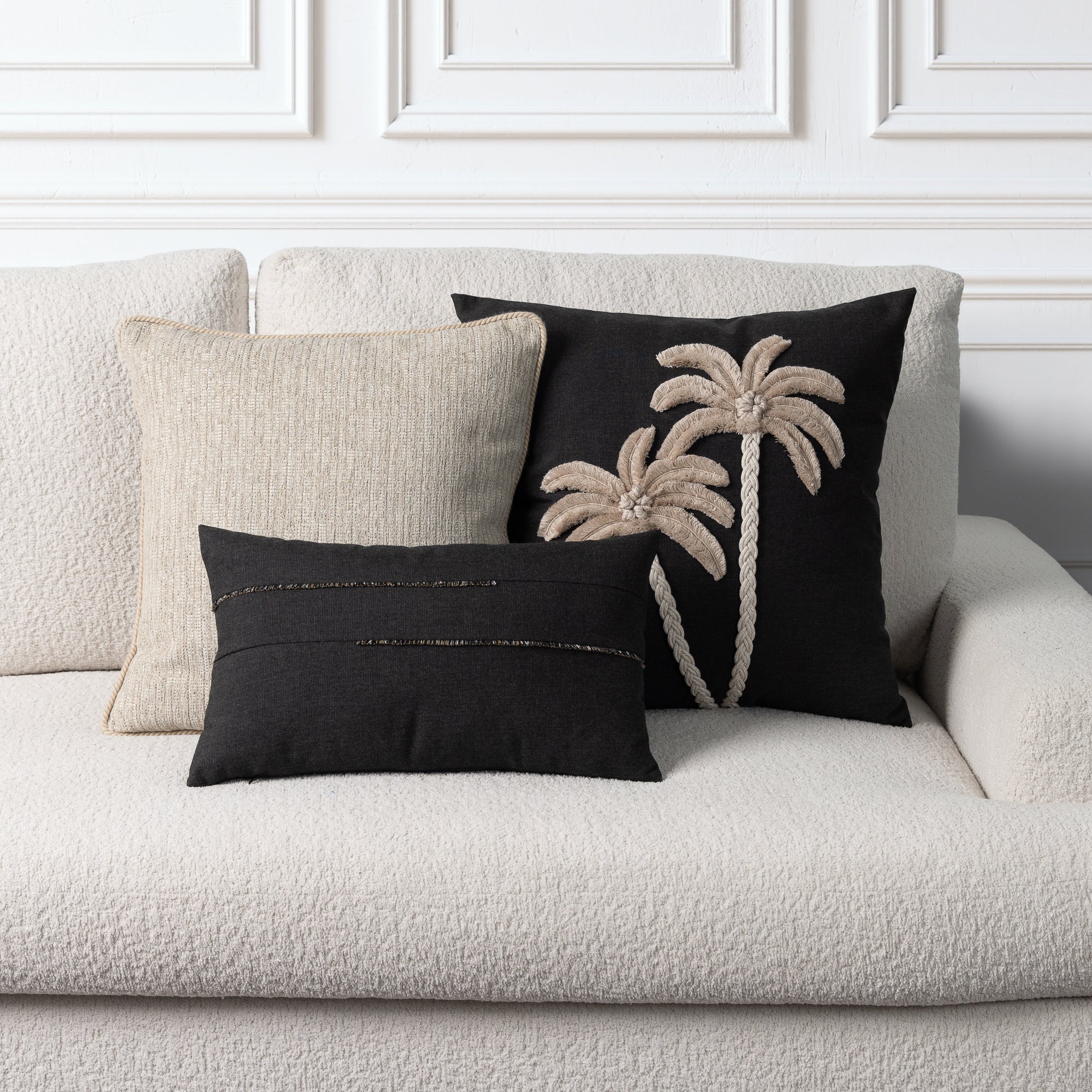 Three decorative throw pillows arranged on a textured cream boucle sofa against a white paneled wall. The set includes a large black pillow with beige rope-style palm tree embroidery, a smaller black pillow with horizontal gold metallic stitching, and a neutral beige woven pillow.