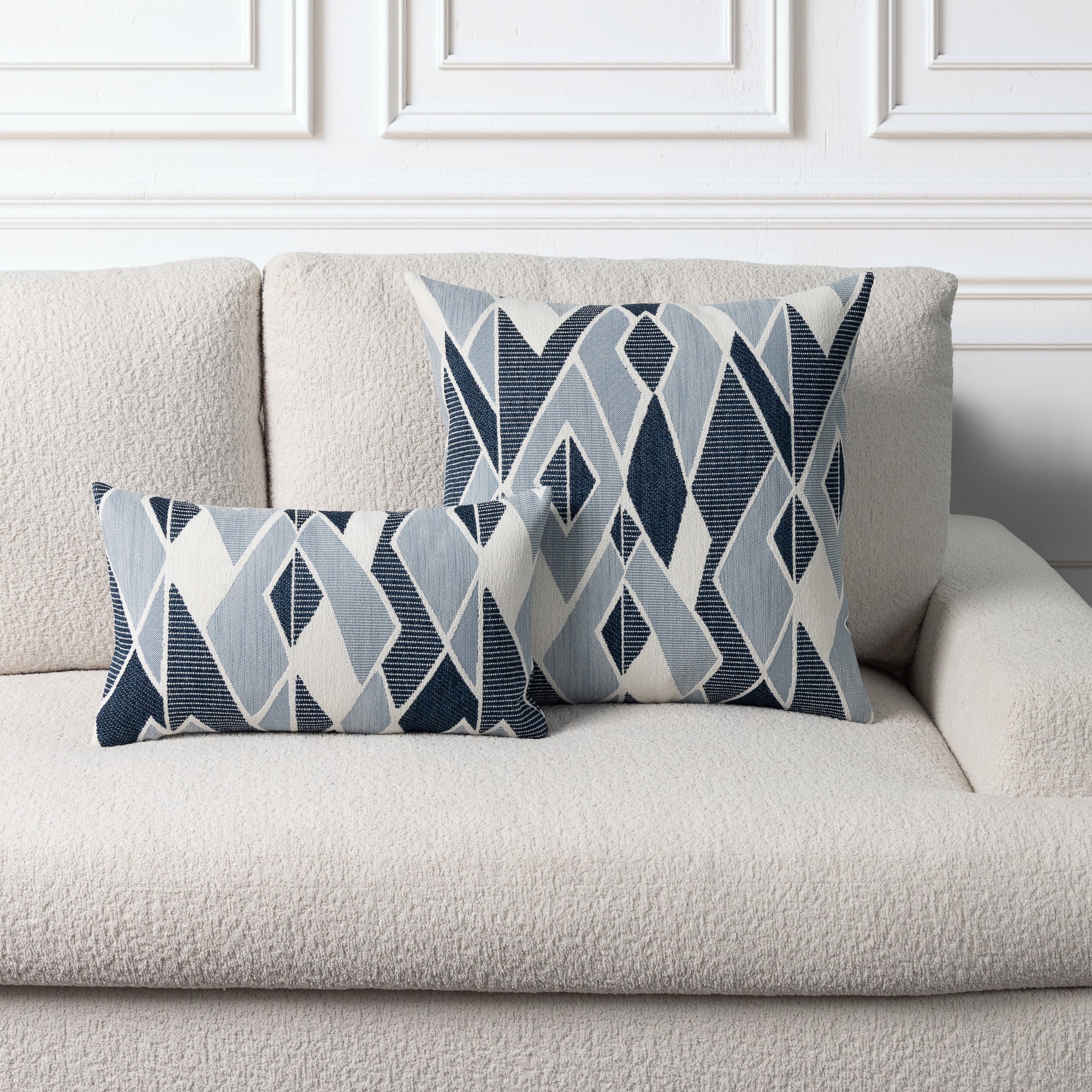 Two decorative throw pillows with a geometric diamond pattern in navy, light blue, and white, styled on a textured cream boucle sofa against a white paneled wall.