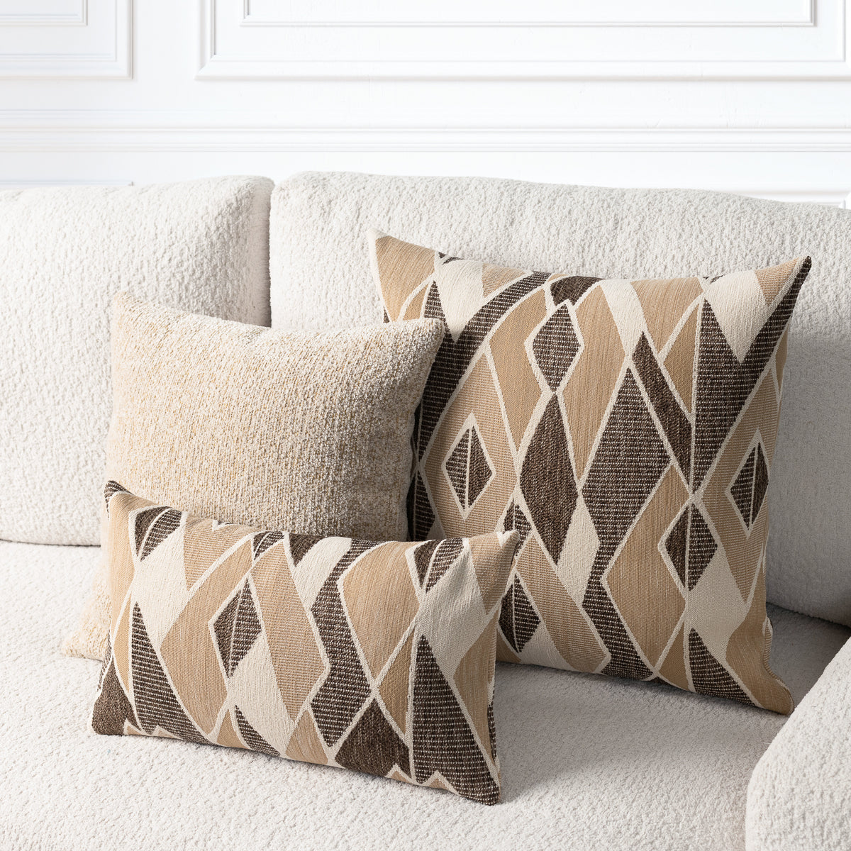 Three decorative throw pillows arranged on a textured cream sofa. Two pillows feature a geometric diamond pattern in beige, brown, and cream, and one pillow is a solid cream color.
