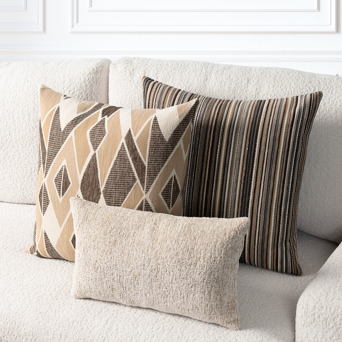 Three decorative throw pillows in neutral tones arranged on a cream textured sofa. The front pillow is a small textured lumbar in beige.