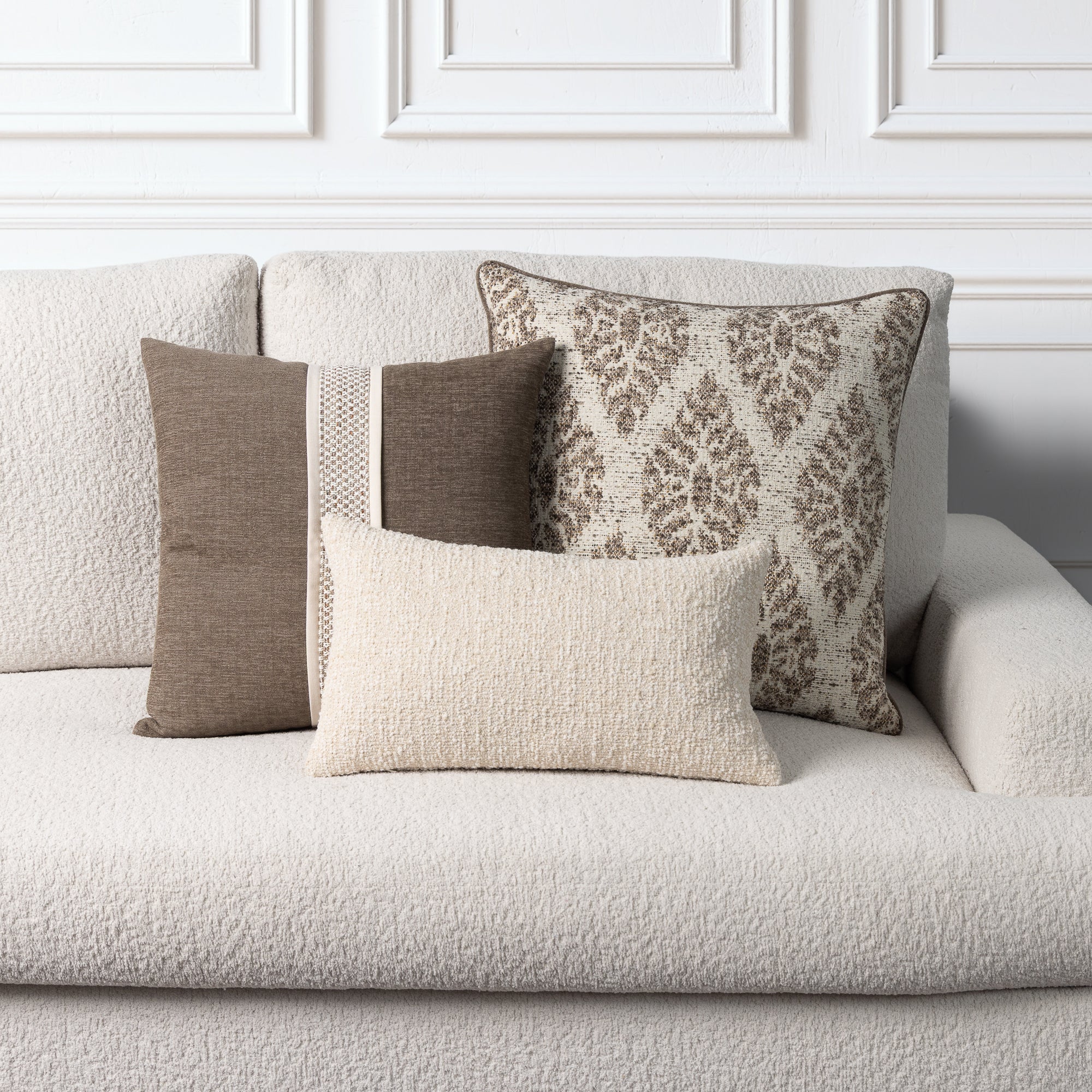 Four decorative throw pillows in neutral tones arranged on a light beige textured sofa against a white paneled wall.
