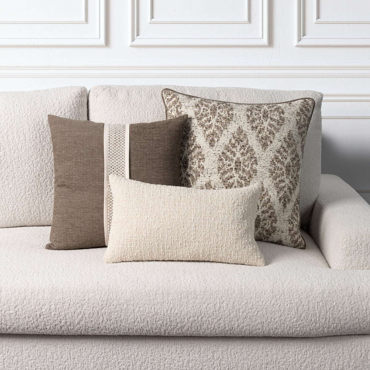Four decorative throw pillows in neutral tones arranged on a light beige textured sofa against a white paneled wall.