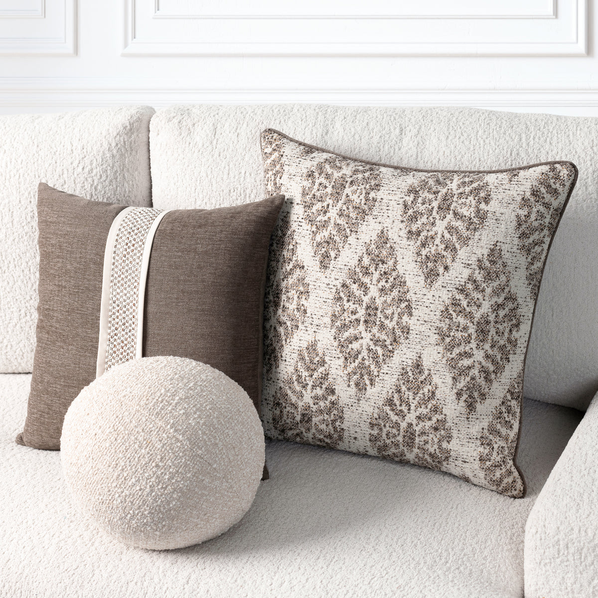 Three decorative throw pillows arranged on a textured cream sofa: a large square pillow with a brown damask pattern on a cream background, a smaller brown pillow with a vertical cream stripe, and a round cream pillow in front.