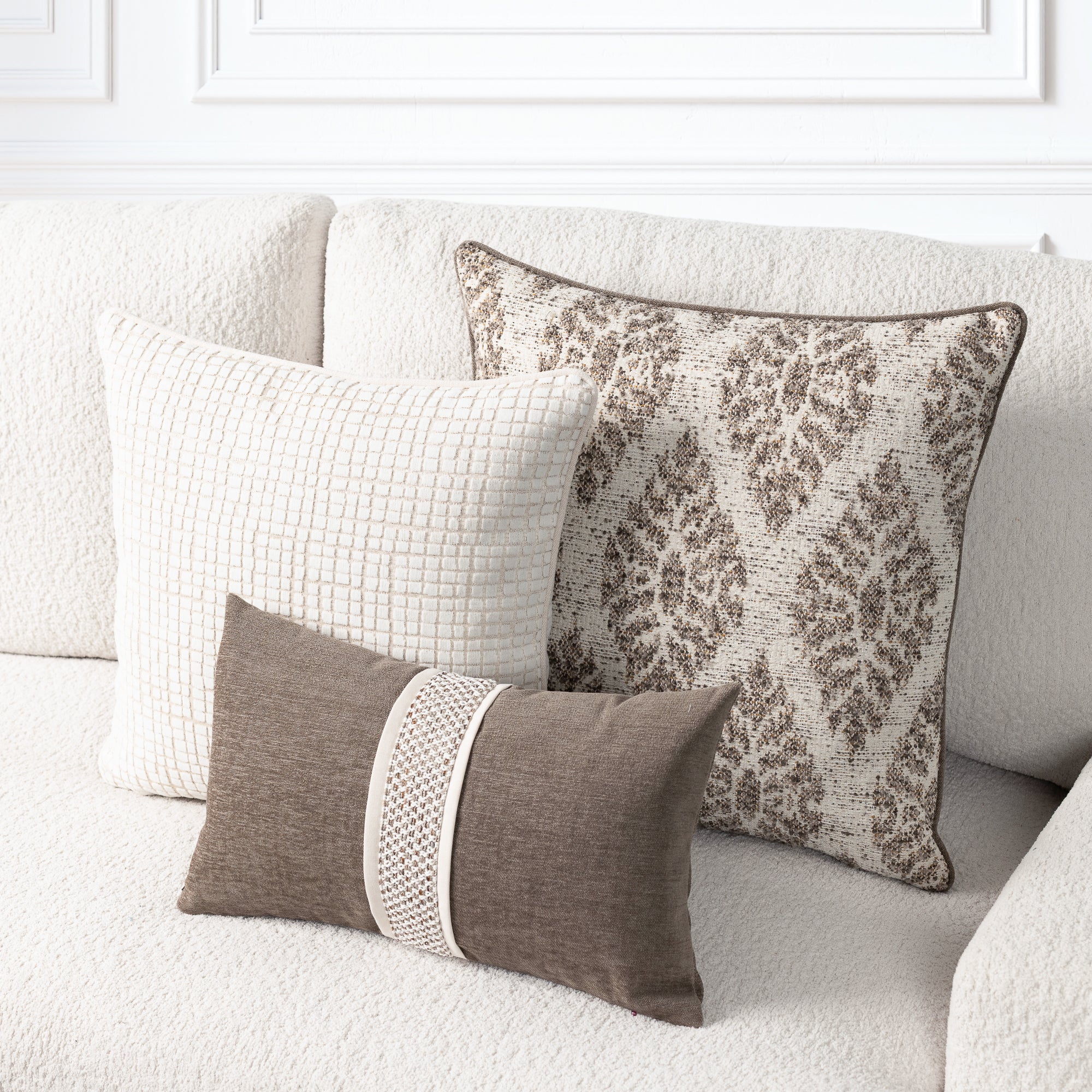 Three decorative throw pillows arranged on a light beige textured sofa. The set includes a small brown pillow with a cream woven center stripe, a larger cream pillow with a subtle grid pattern, and a larger brown-and-cream damask-patterned pillow.