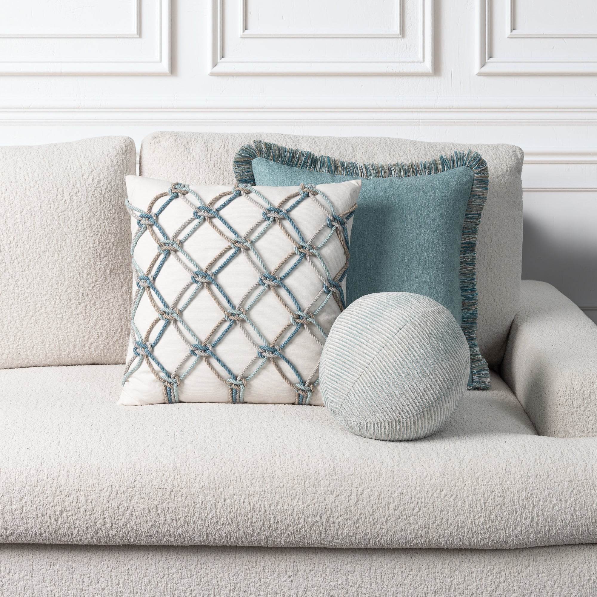 Three decorative throw pillows arranged on a light beige textured sofa against a white paneled wall. The pillows include a square cream pillow with a blue and taupe geometric lattice pattern, a solid dusty blue square pillow with fringe trim, and a round ribbed knit pillow in a light blue-gray.