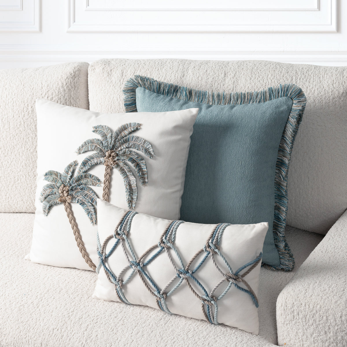 Three decorative throw pillows arranged on a textured cream sofa: a square pillow with embroidered palm trees, a teal fringed pillow, and a rectangular cream pillow with a rope lattice pattern.