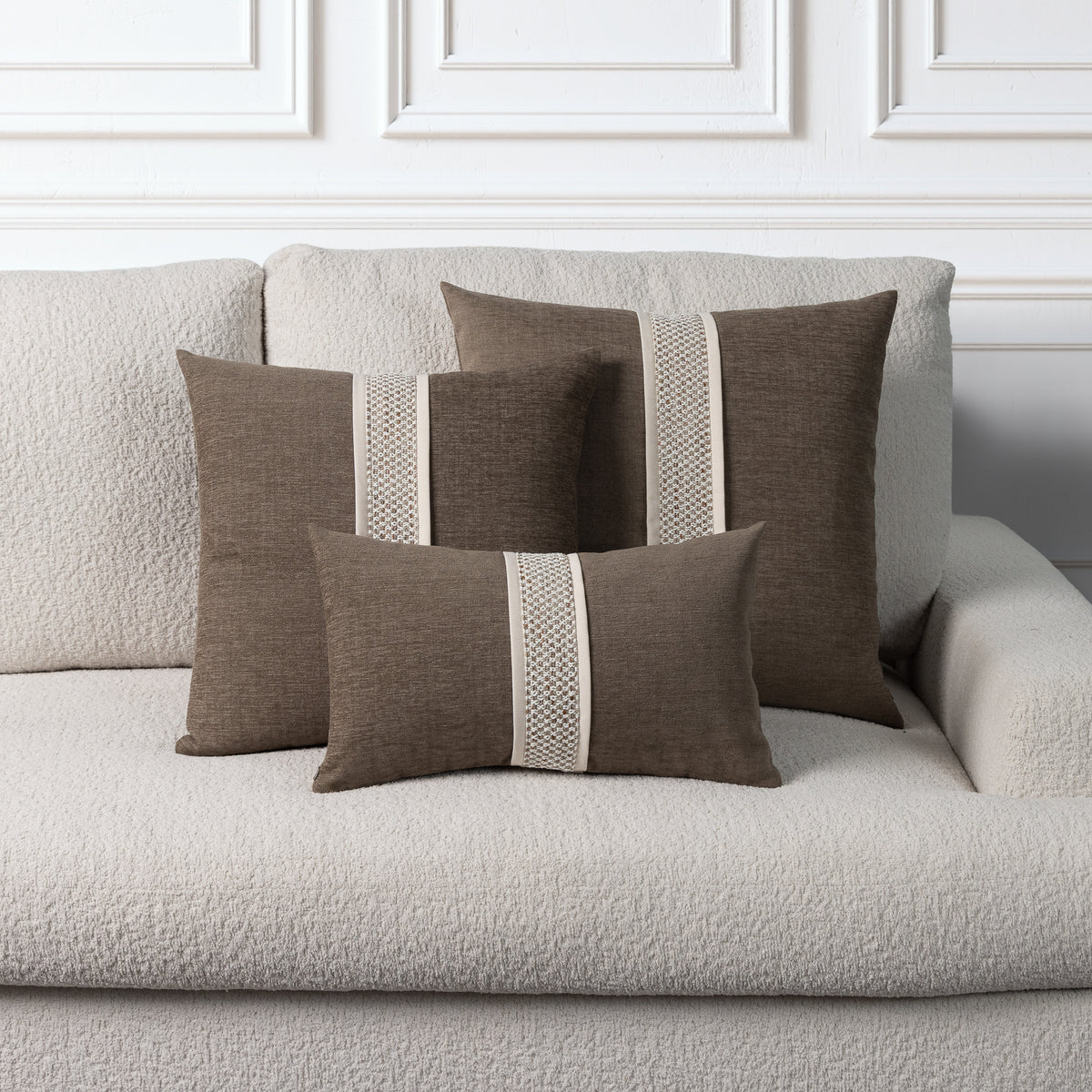 Four brown throw pillows with vertical cream textured stripe accents, arranged on a light beige boucle sofa against a white paneled wall.