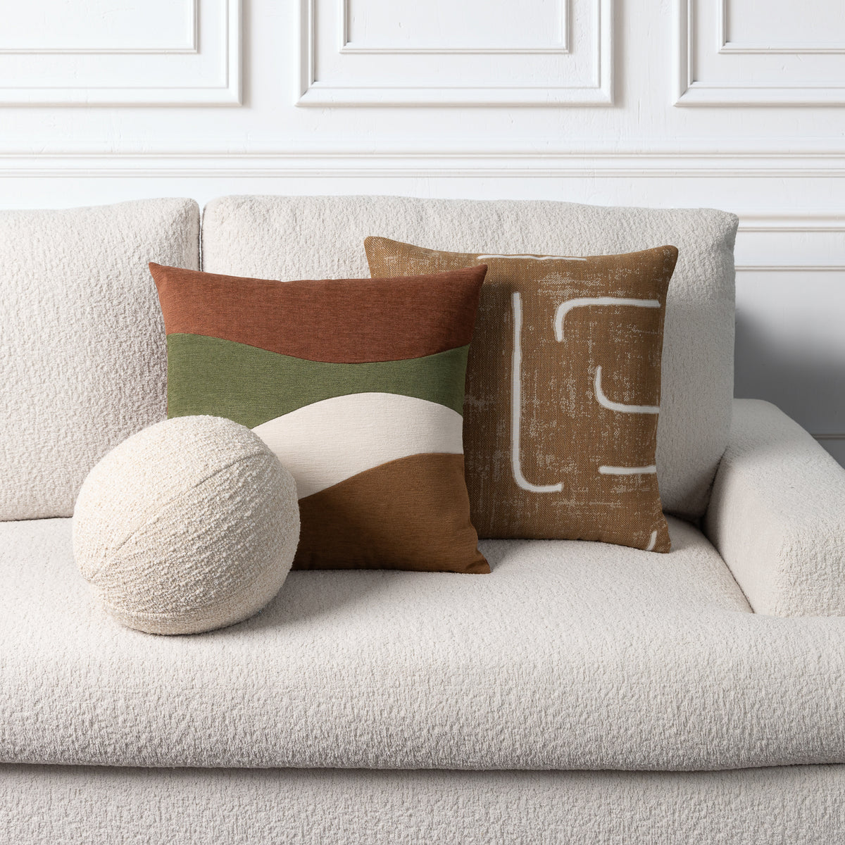 Three decorative throw pillows arranged on a textured off-white sofa against a white paneled wall. The set includes a round cream pillow, a square pillow with horizontal terracotta, olive green, and cream bands, and a square tan pillow with a white abstract line pattern.