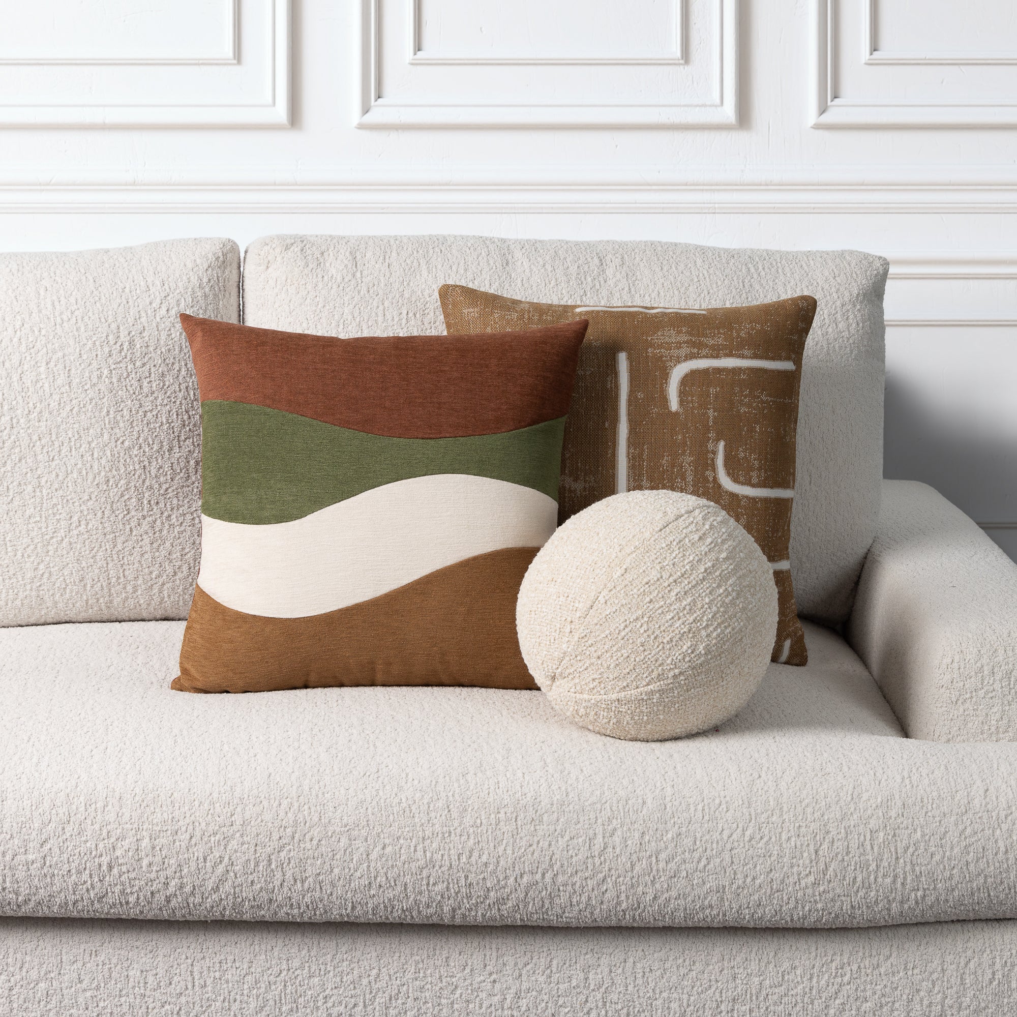 A styled interior scene showing two decorative throw pillows and a round textured pillow on a light beige boucle sofa against a white paneled wall. The front pillow has broad horizontal bands in rust, olive green, cream, and tan, while the rear pillow has a tan abstract pattern.
