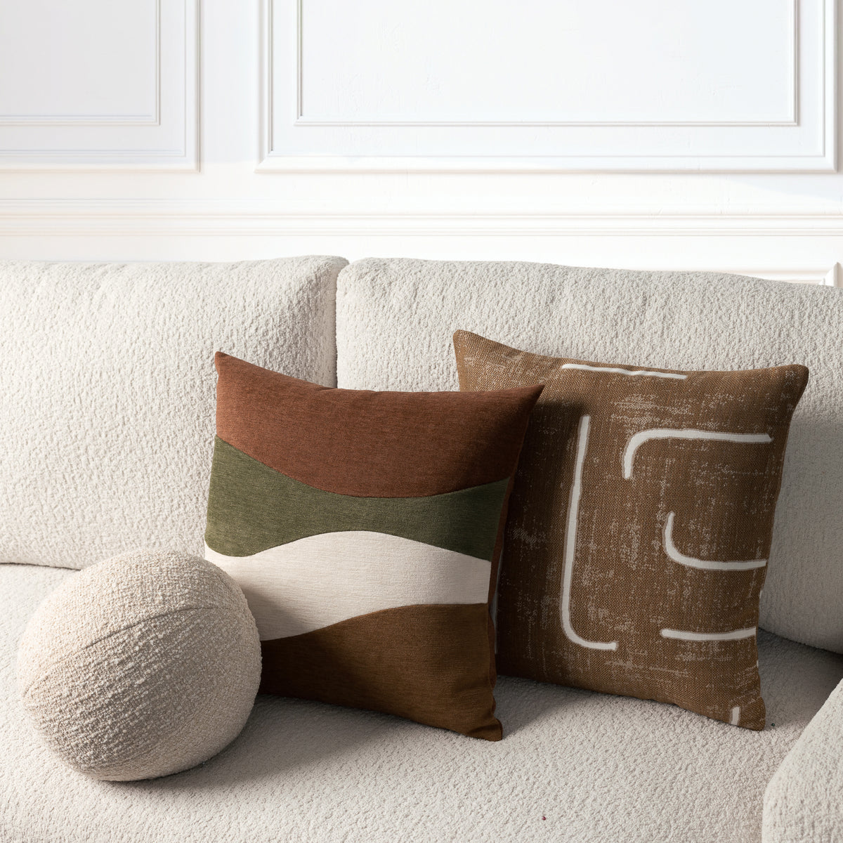 Two decorative throw pillows and a round textured pillow on a light beige sofa against a white paneled wall. One pillow has horizontal bands in rust, olive green, cream, and brown, while the other has a brown abstract white line pattern.