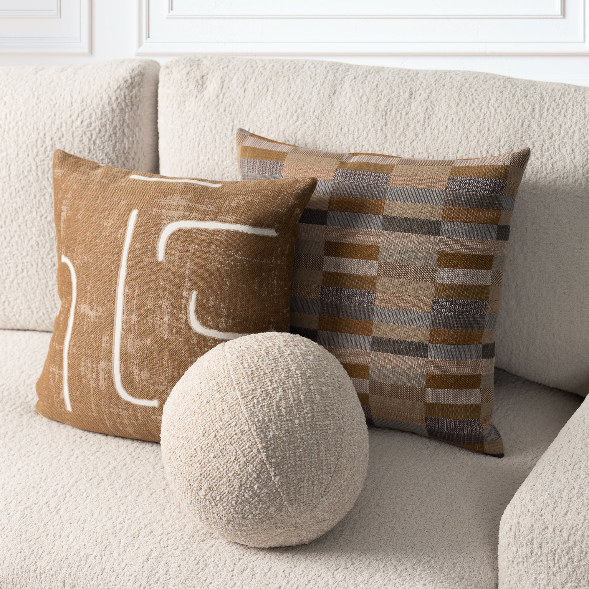 Three decorative throw pillows arranged on a textured cream sofa: a square tan pillow with white abstract line art, a square plaid pillow in tan, brown, and gray, and a round cream textured pillow in front.