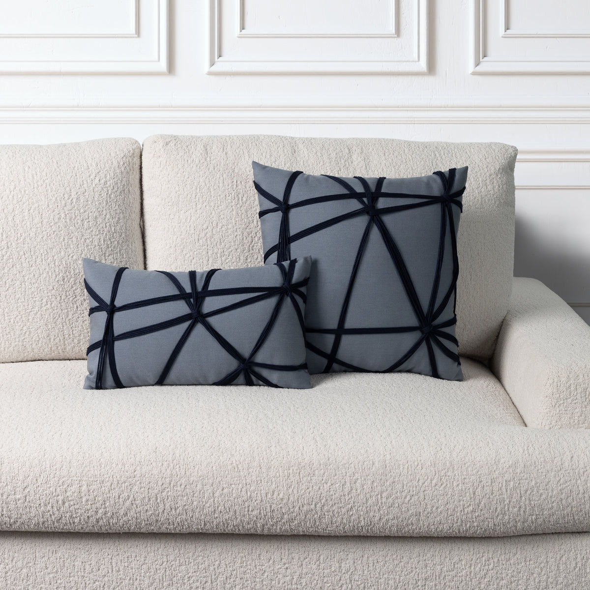 Two square throw pillows with a dark navy geometric line pattern on a slate gray background, styled on a cream bouclé sofa against a white paneled wall.