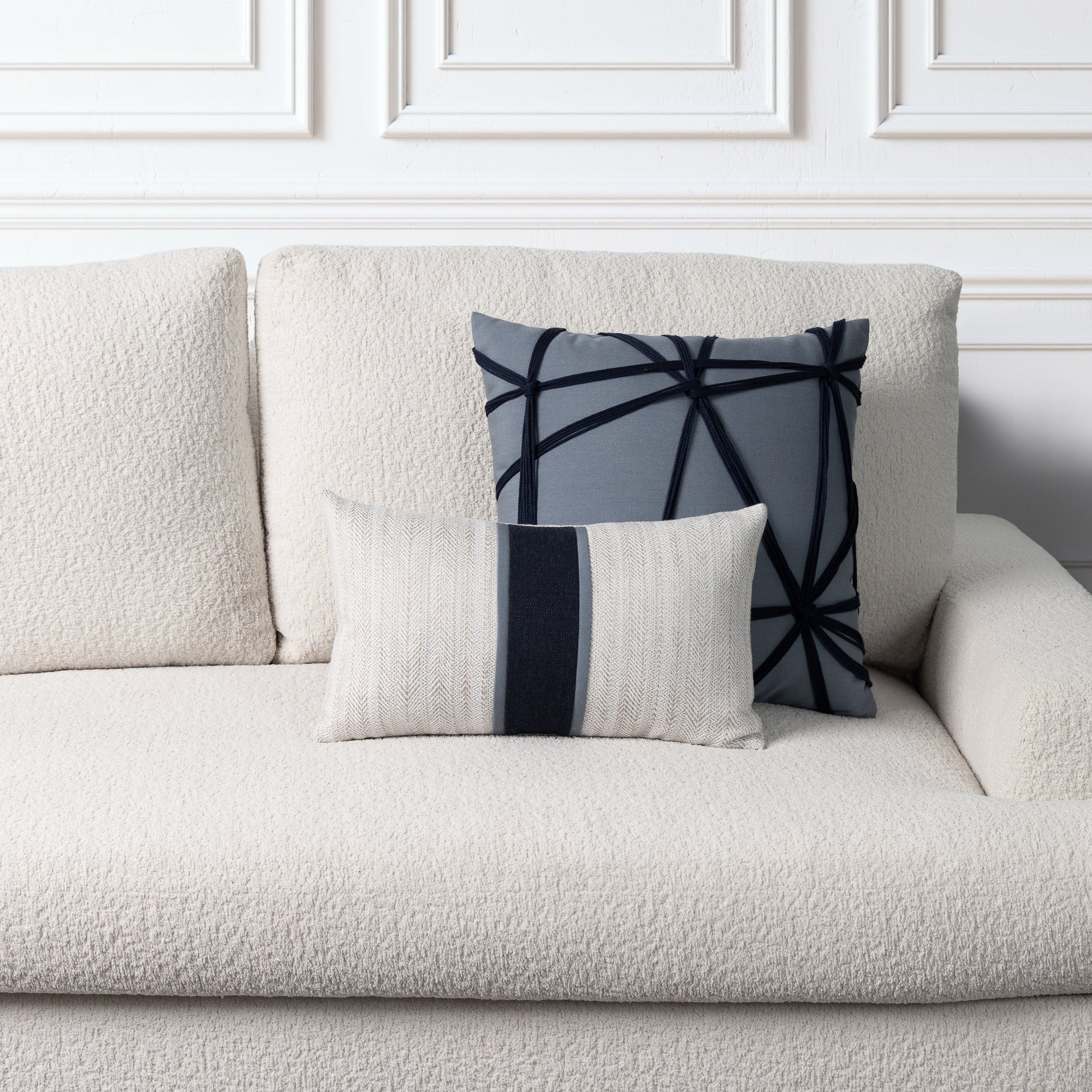 Two decorative throw pillows arranged on a textured cream boucle sofa against a white paneled wall. The pillows include a gray one with indigo geometric lines, and a cream one with a vertical indigo stripe.