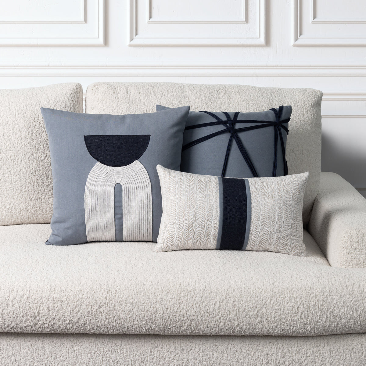 Three decorative throw pillows arranged on a textured cream boucle sofa against a white paneled wall. The pillows include a slate blue one with a white and indigo arch motif, a gray one with indigo geometric lines, and a cream one with a vertical indigo stripe.