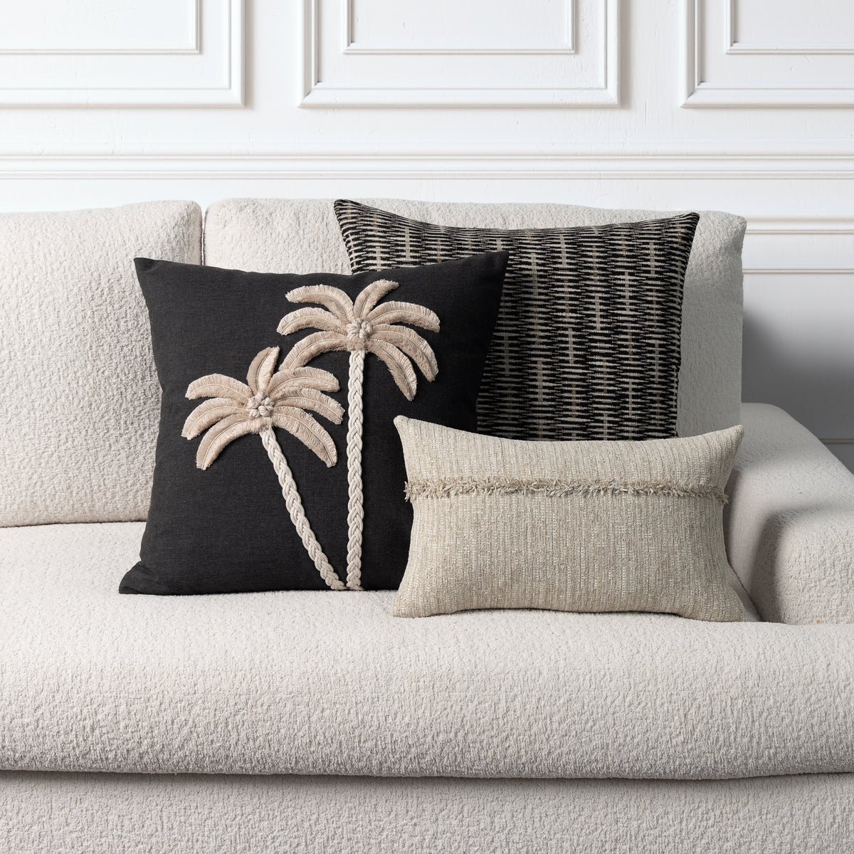 Three decorative throw pillows arranged on a light beige textured sofa against a white paneled wall. The left pillow is black with beige palm tree embroidery; the center pillow is black and white woven; the front pillow is light beige with fringe trim.