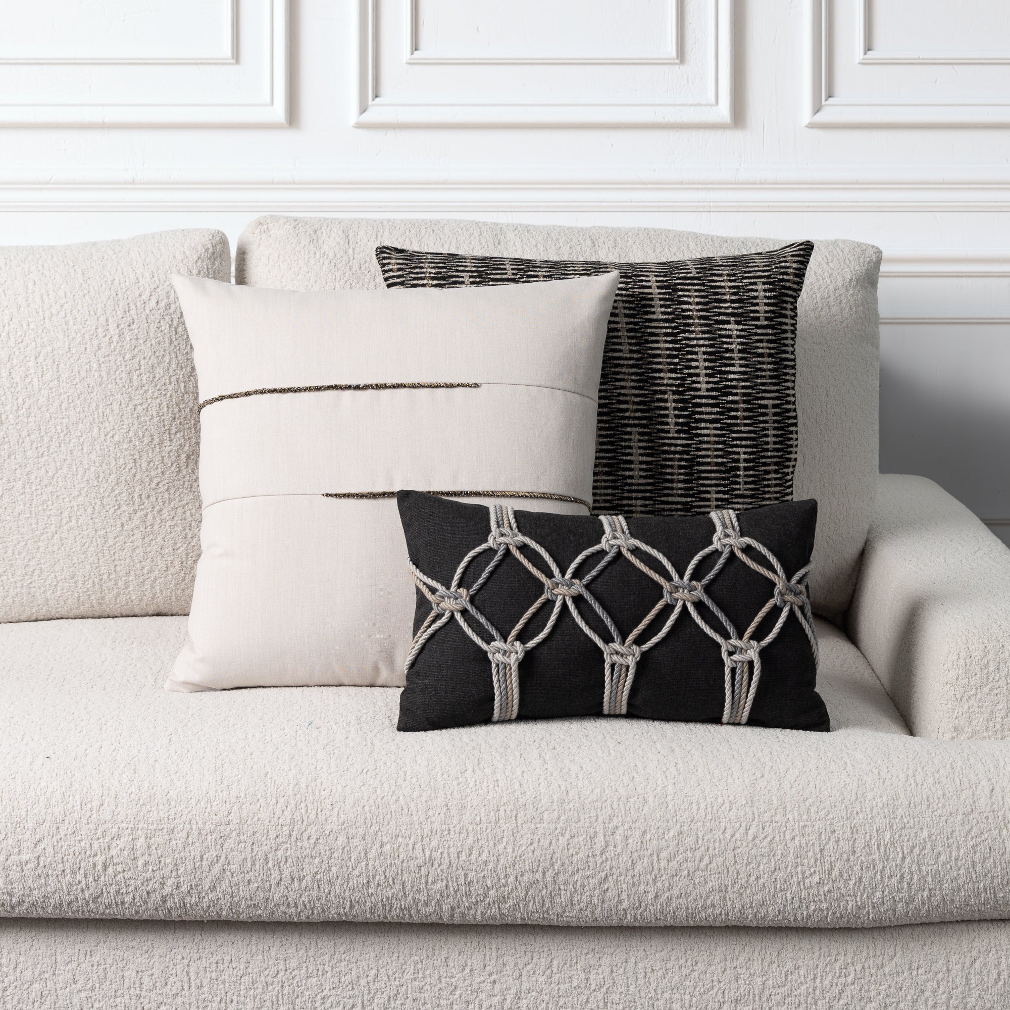 Three decorative throw pillows arranged on a textured light beige sofa against a white paneled wall. The pillows include a large cream pillow with a thin dark stripe, a black-and-white woven vertical pillow, and a black lumbar pillow with a cream geometric knot pattern.