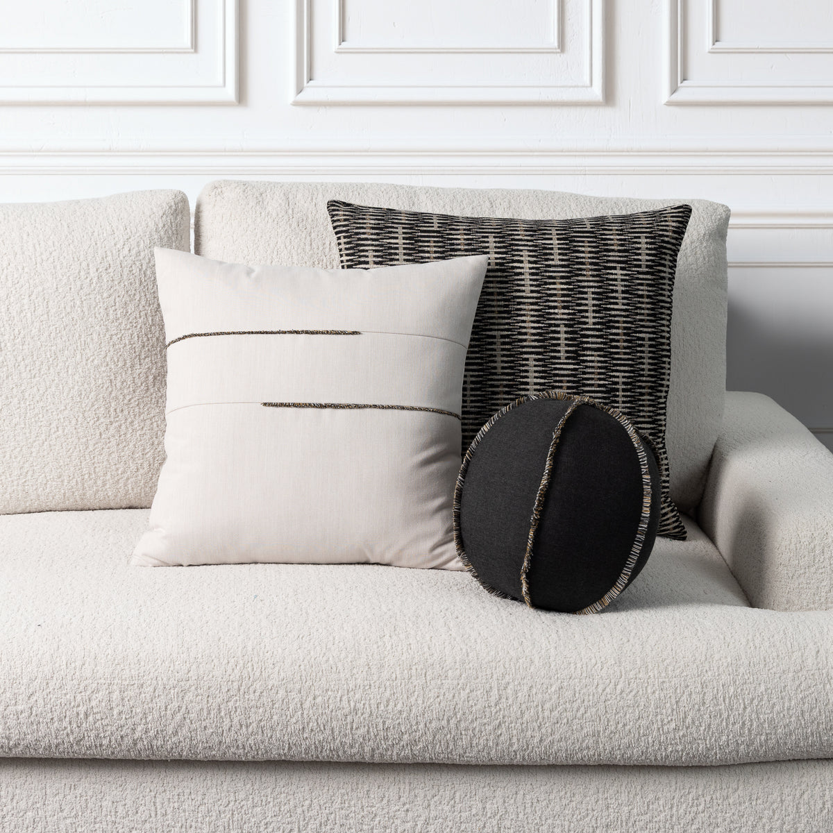 Three decorative throw pillows arranged on a light beige textured sofa against a white paneled wall. The set includes a large cream pillow with two horizontal black stitched lines, a black and gray woven patterned pillow, and a small round black lumbar pillow with a fringed edge.
