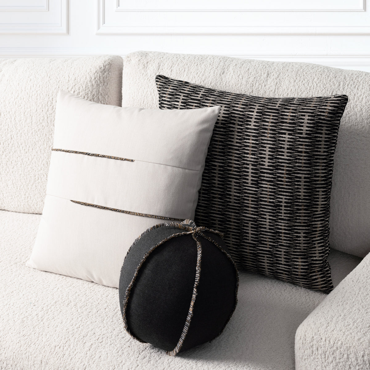 Three decorative throw pillows arranged on a textured off-white sofa: a cream pillow with two horizontal metallic trim lines, a black-and-cream woven pattern pillow, and a round black pillow with metallic trim.