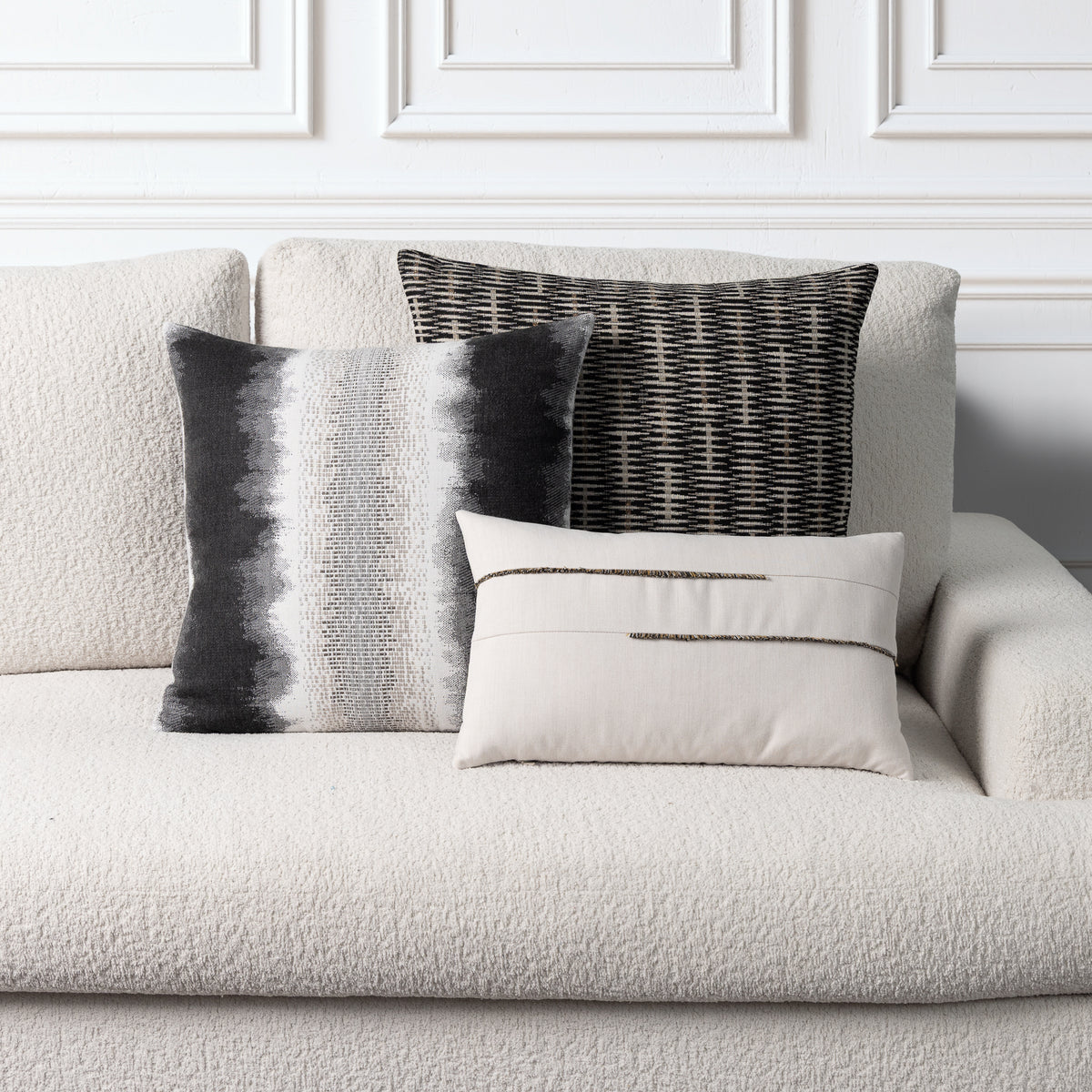 Three decorative throw pillows styled on a textured off-white boucle sofa against a white paneled wall. The pillows include a square black-and-white striped pillow, a black-and-white woven pillow, and a smaller cream pillow with two horizontal black stripes.
