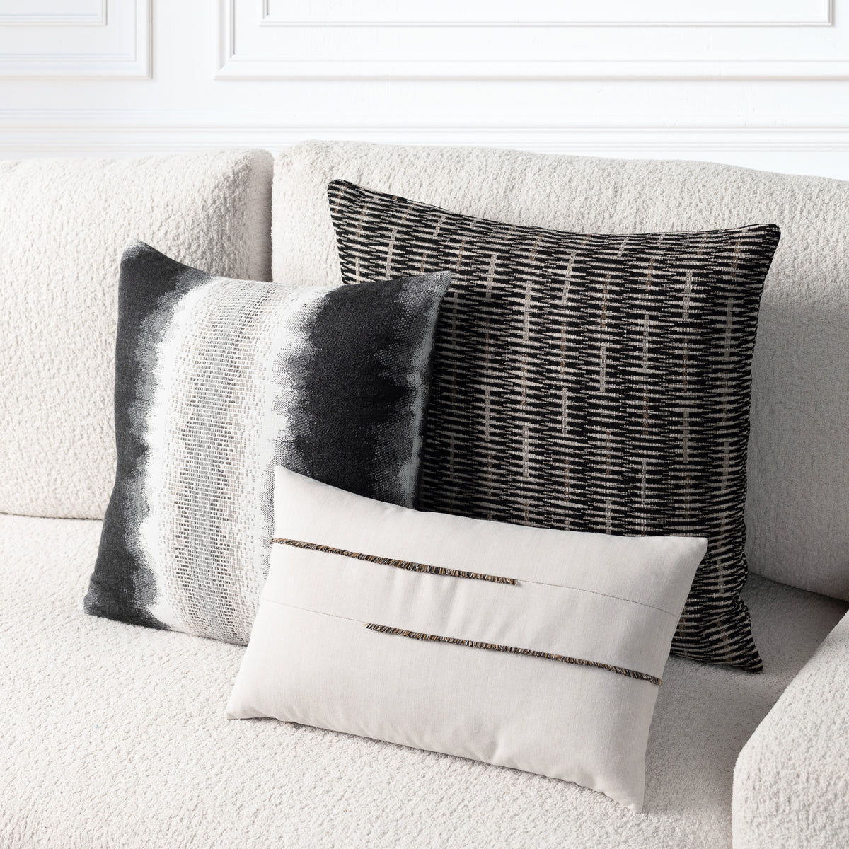 Three decorative throw pillows arranged on a textured cream sofa. The set includes a square black-and-white striped pillow, a black-and-white woven pattern pillow, and a smaller rectangular cream pillow with two horizontal metallic trim details.