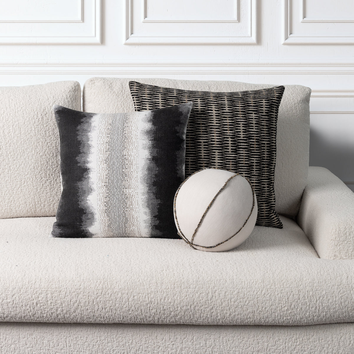 Three decorative throw pillows arranged on a light beige textured sofa against a white paneled wall. One square pillow has vertical black, gray, and white stripes, one square pillow has a black-and-white woven pattern, and one round pillow is off-white with dark stitched lines.