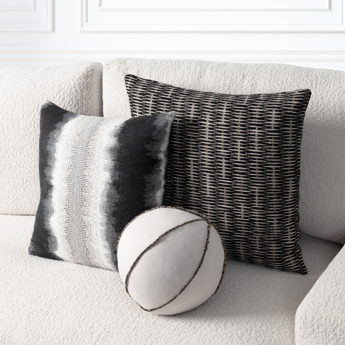 Three decorative throw pillows arranged on a textured light beige sofa: a square pillow with vertical gray and white stripes, a square pillow with a black and beige basketweave pattern, and a round pillow in off-white with dark contrast stitching.