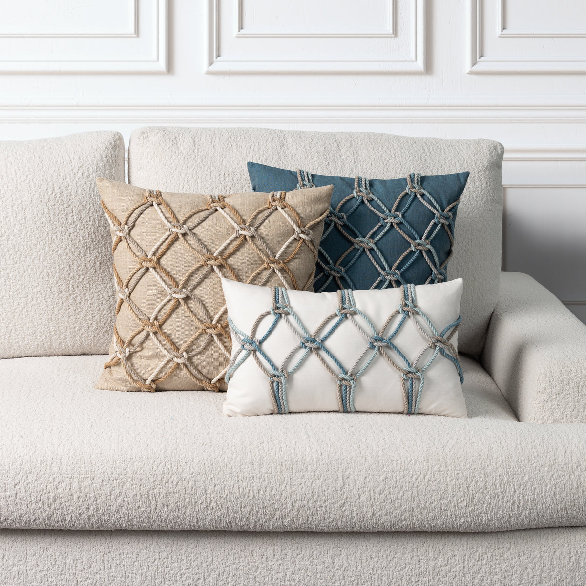 Three decorative throw pillows with geometric rope-knit patterns arranged on a textured light beige sofa against a white paneled wall.