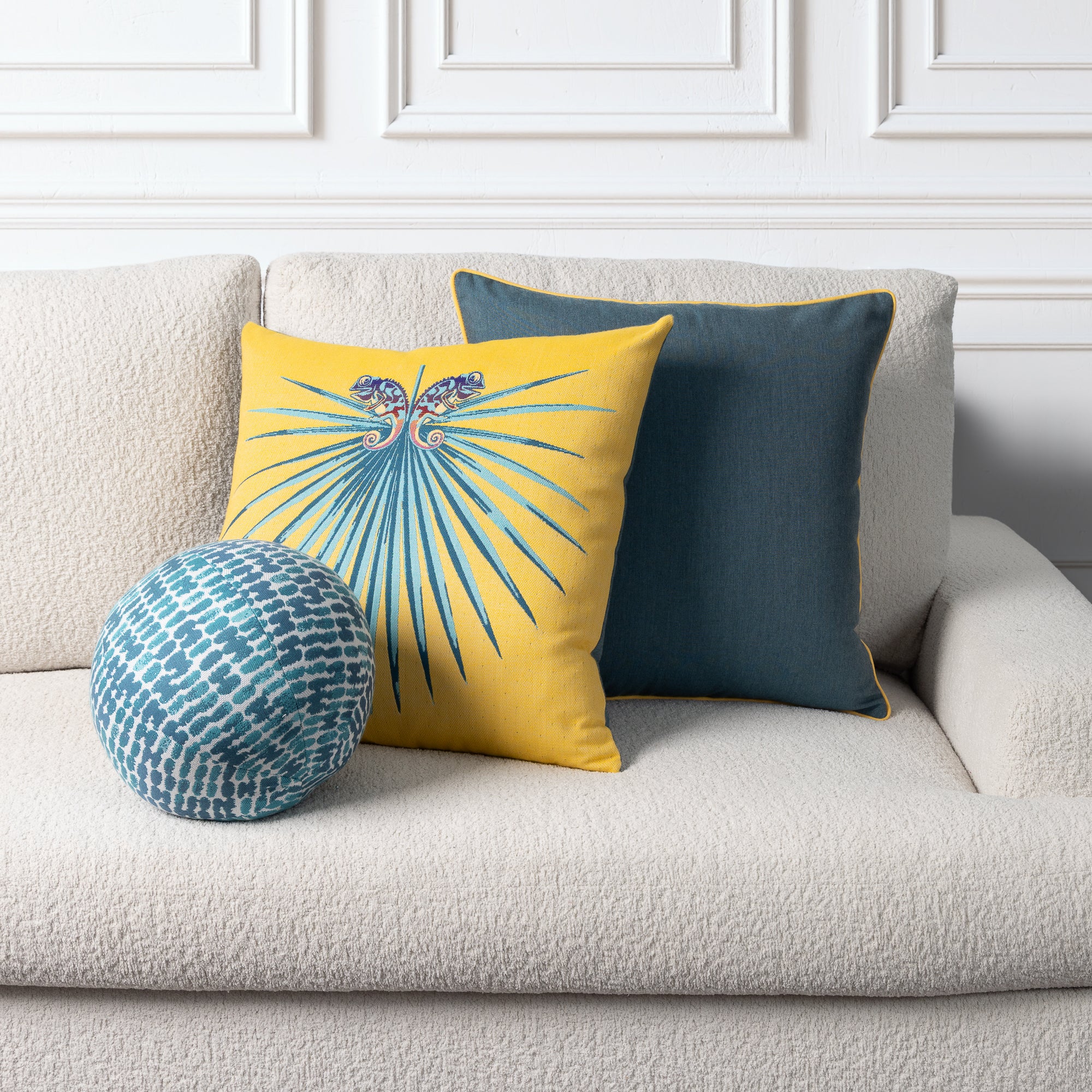 Three decorative throw pillows arranged on a light beige textured sofa: a round blue-and-white patterned pillow, a square yellow pillow with a blue sunburst and butterfly motif, and a square dark teal pillow with a subtle sheen.