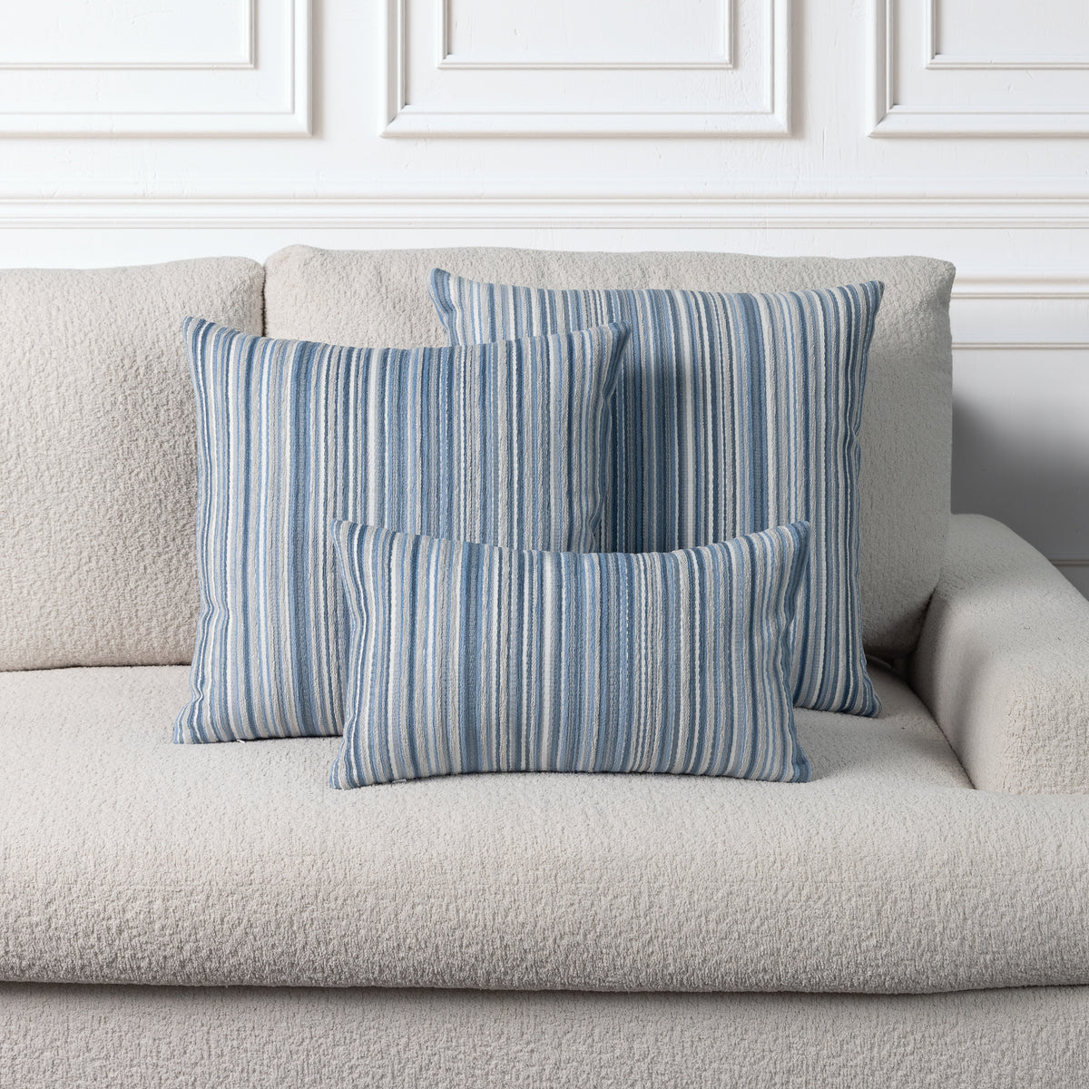Three striped throw pillows in shades of blue, navy, and white, arranged on a textured light beige sofa against a white paneled wall.