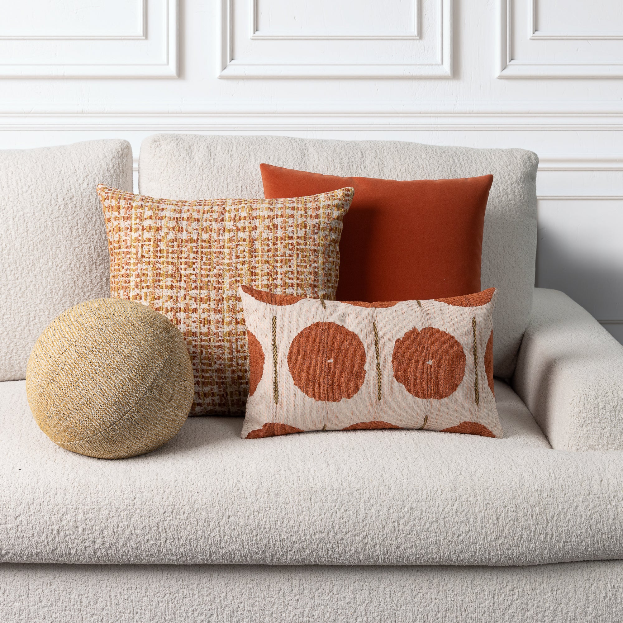 Three decorative throw pillows styled on a textured off-white boucle sofa against a white paneled wall. The set includes a round natural jute pillow, a cream pillow with a rust-orange basketweave pattern, a solid rust-orange square pillow, and a cream lumbar pillow with two large rust-orange circular motifs.