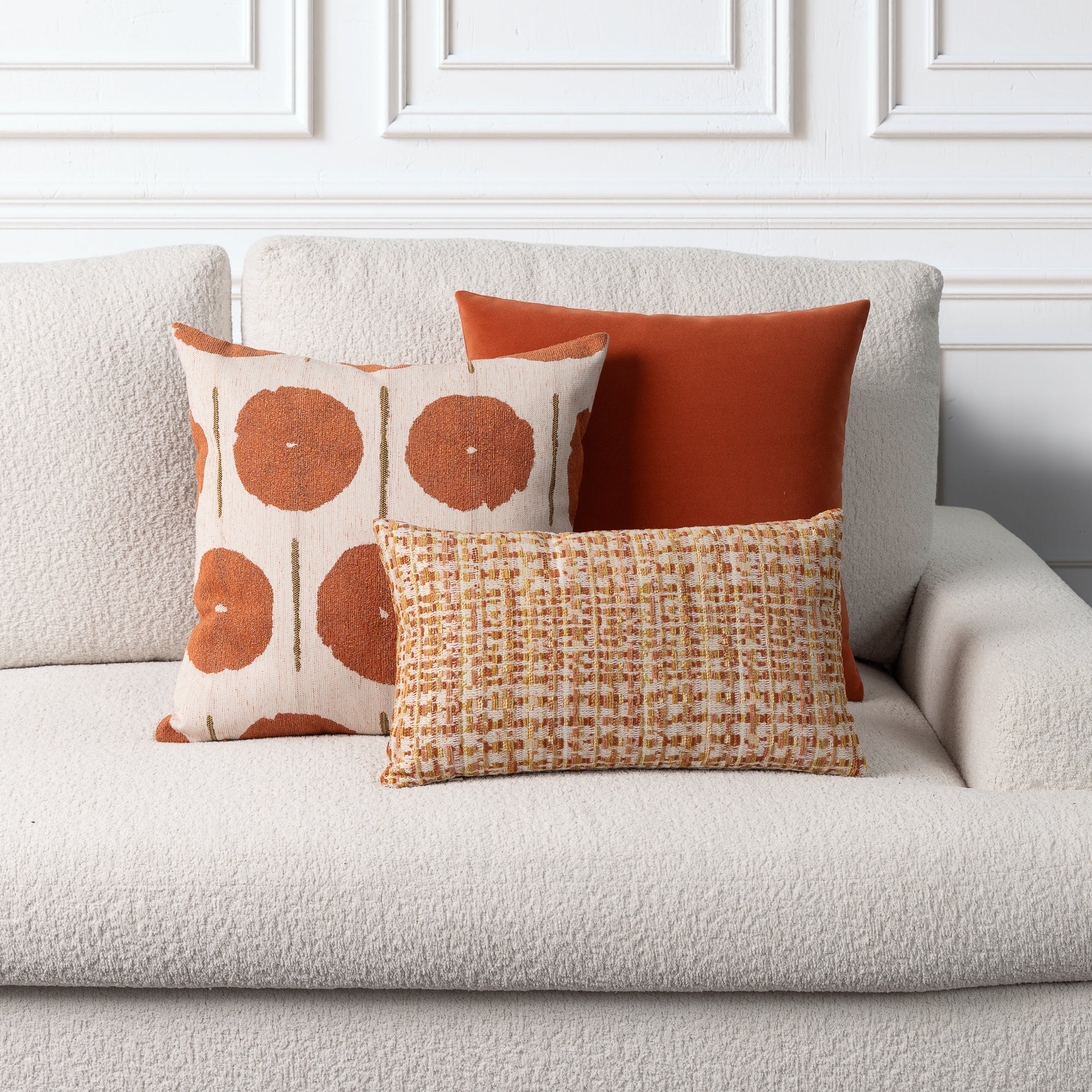 Three decorative throw pillows arranged on a textured off-white sofa against a white paneled wall. The pillows include a cream pillow with large rust-orange circular motifs, a solid rust-orange pillow, and a smaller woven rectangular pillow in a warm tan and orange pattern.
