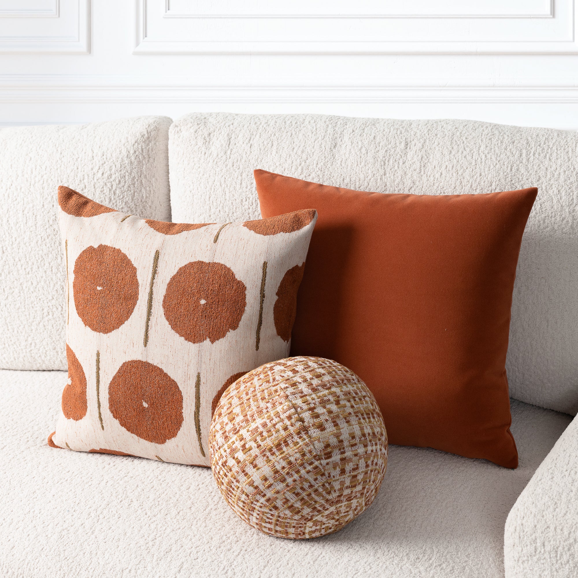 Three decorative throw pillows arranged on a textured cream boucle sofa: a square pillow with a rust-orange circular dot pattern on a cream background, a solid rust-orange square pillow, and a round woven pillow in a tan-and-cream basketweave pattern.