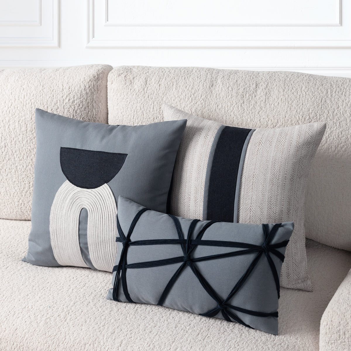 Three decorative throw pillows in gray, beige, and indigo tones arranged on a textured cream sofa. The pillows feature abstract geometric and striped patterns.