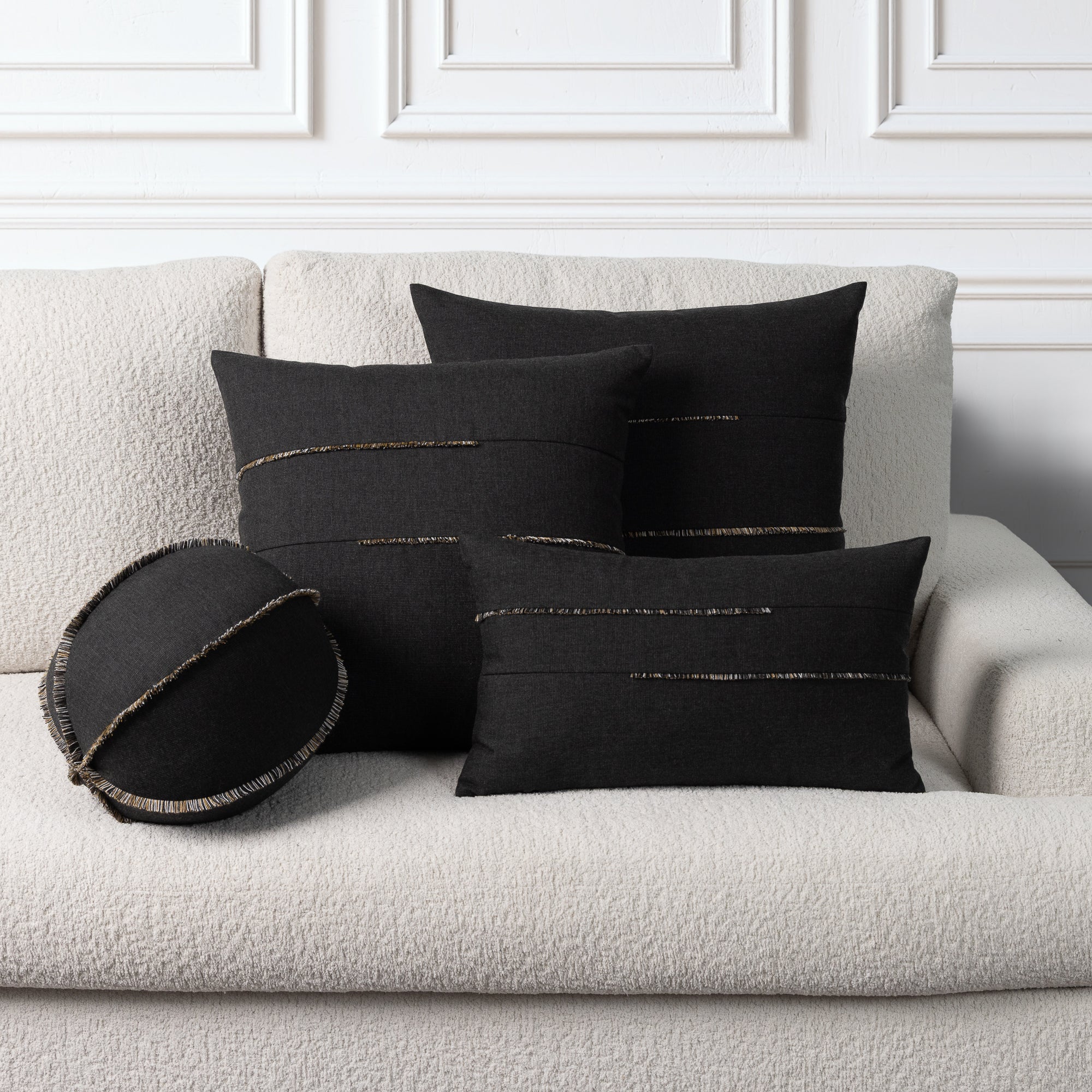 Four black decorative throw pillows with metallic gold stitched details arranged on a textured off-white boucle sofa against a white paneled wall.