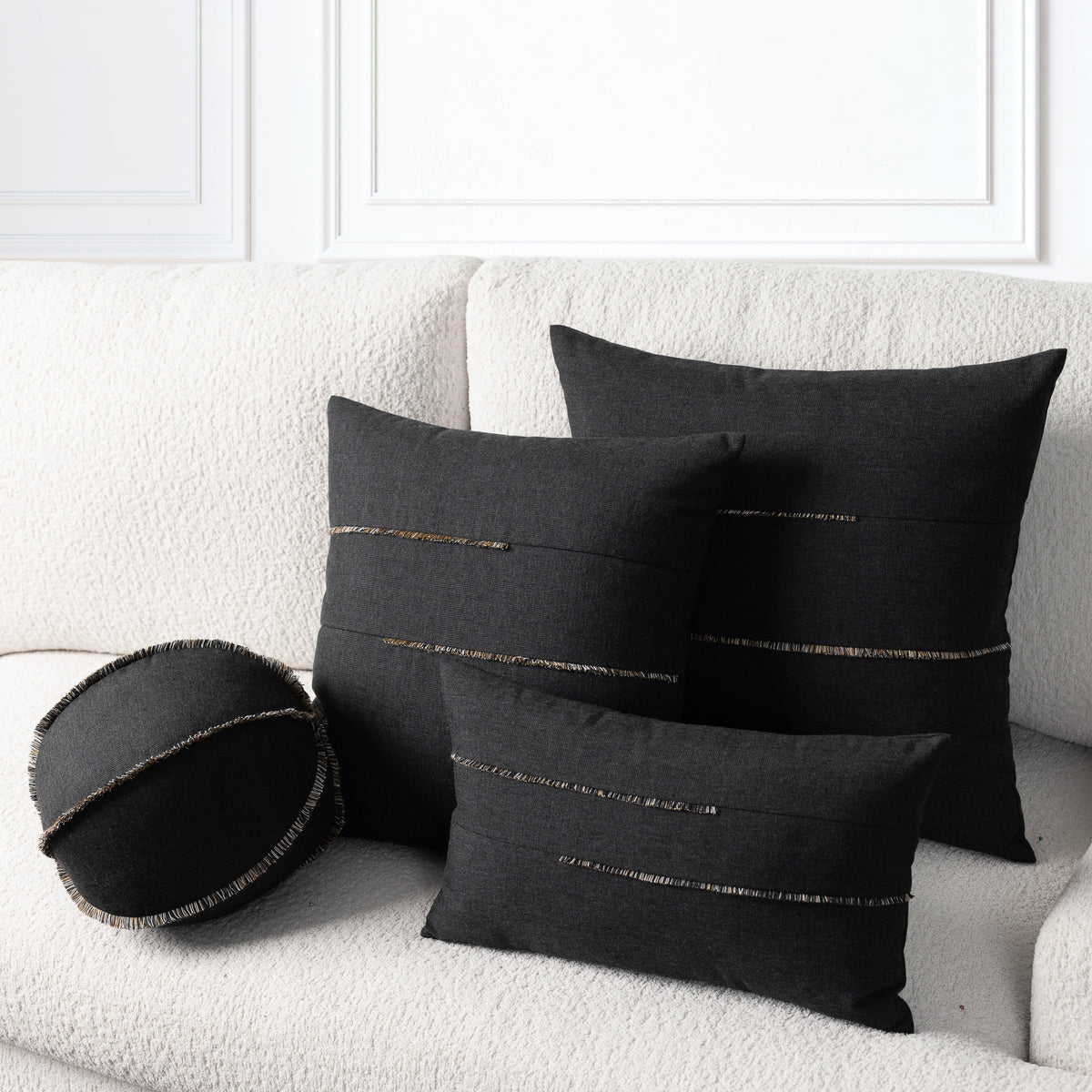 Three black throw pillows with brown and gold horizontal fringe, including two square pillows and one round lumbar pillow, arranged on a textured cream boucle sofa.