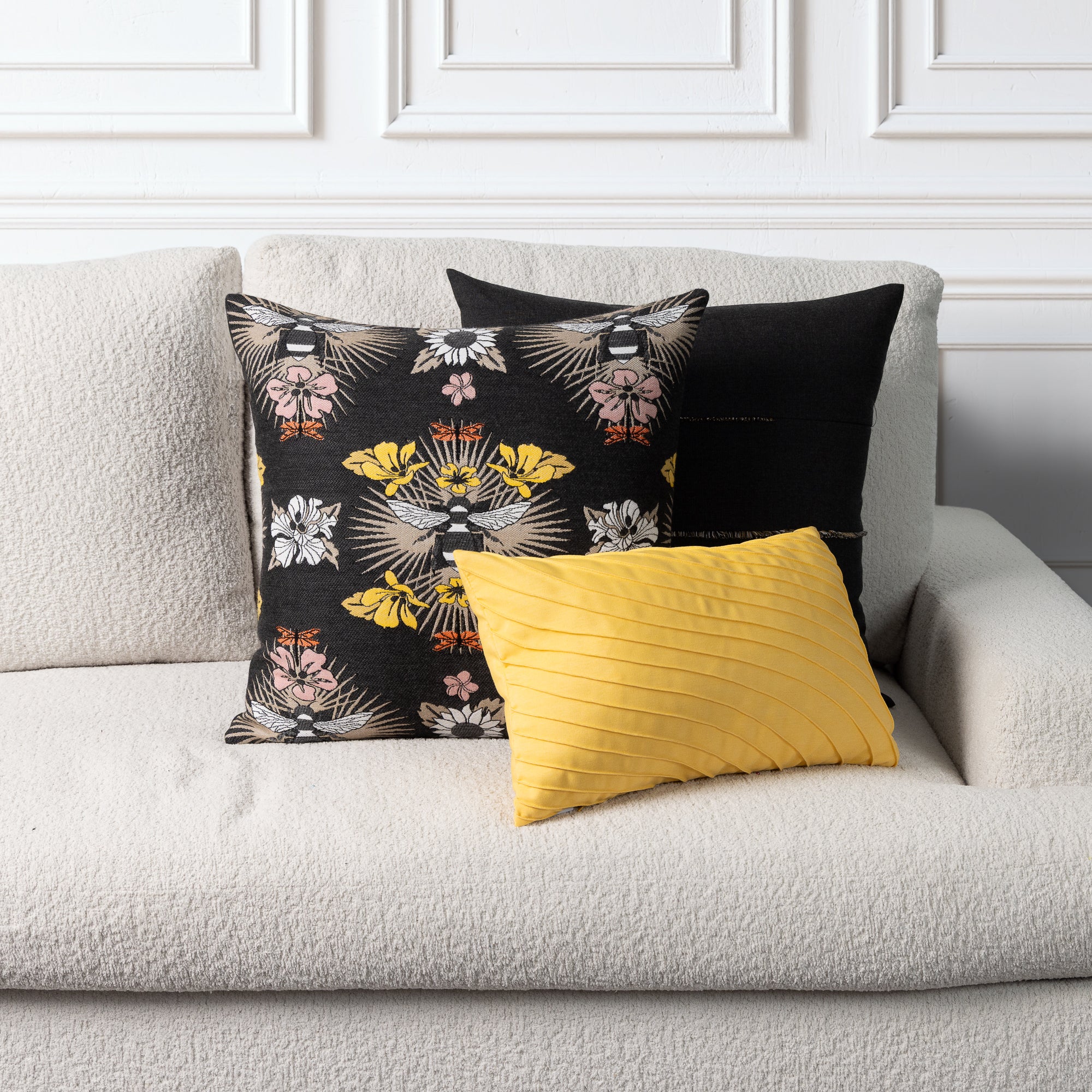 Three decorative throw pillows arranged on a light beige textured sofa against a white paneled wall. The front pillow is mustard yellow with vertical pleats, the left pillow is black with a multicolor floral and bird pattern, and the back pillow is solid black.