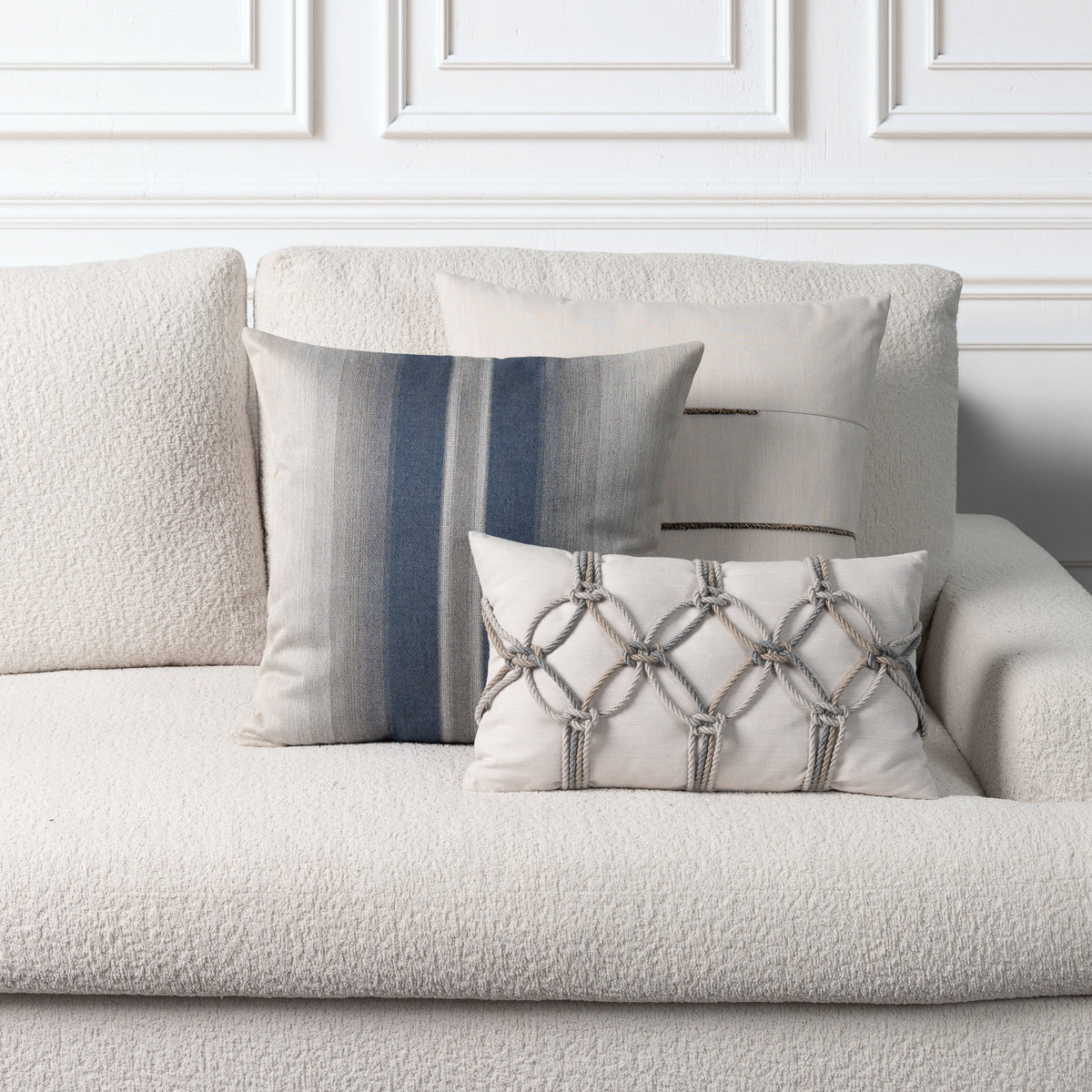Three decorative throw pillows arranged on a textured cream boucle sofa against a white paneled wall. The pillows include a large cream pillow with an indigo vertical stripe pattern, a cream pillow with a horizontal striped fringe decoration, and a cream lumbar pillow with a gray rope-knot geometric pattern.