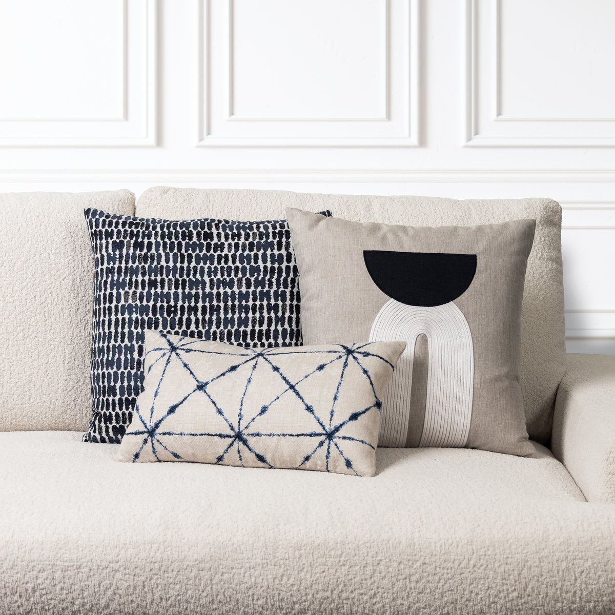 Three decorative throw pillows arranged on a textured cream sofa against a white paneled wall. The pillows include a navy and white geometric pattern, a taupe pillow with a black and white abstract design, and a smaller taupe pillow with a navy geometric lattice pattern.