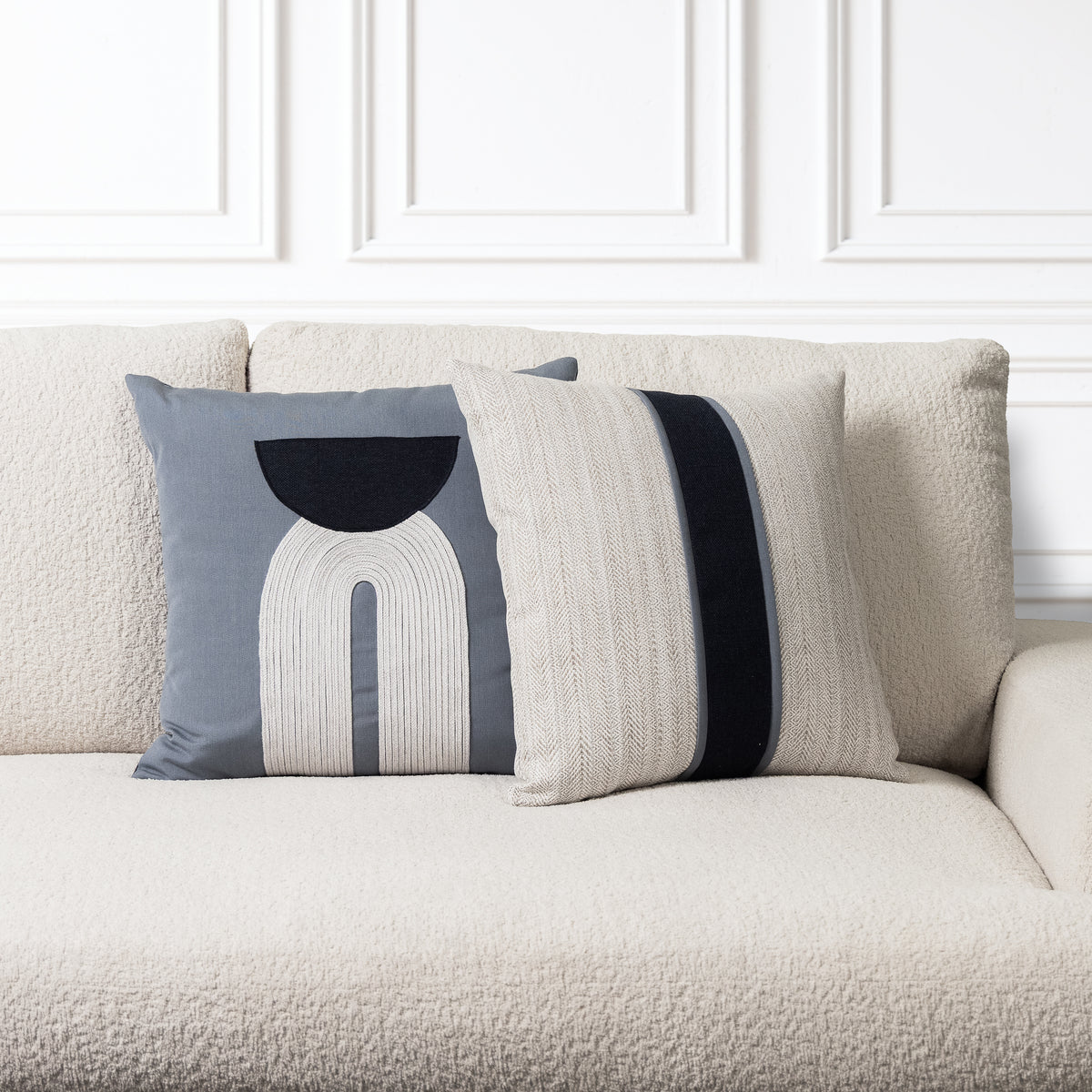 Two decorative throw pillows on a textured cream sofa against a white paneled wall. One pillow is slate blue with a white and black geometric arch design, and the other is cream with a wide black stripe.