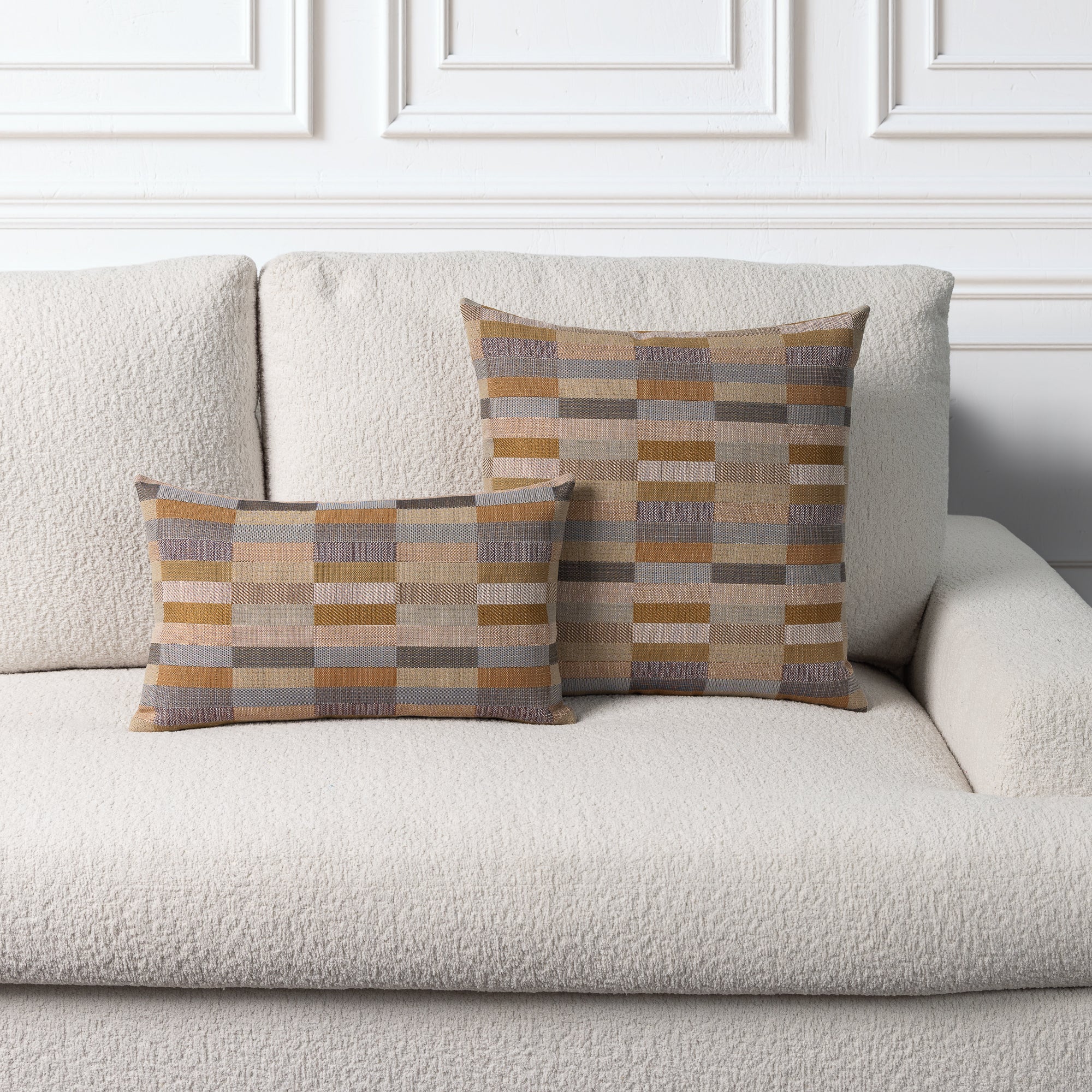 Two decorative throw pillows with a geometric plaid pattern in beige, brown, and gray, styled on a cream boucle sofa against a white paneled wall.
