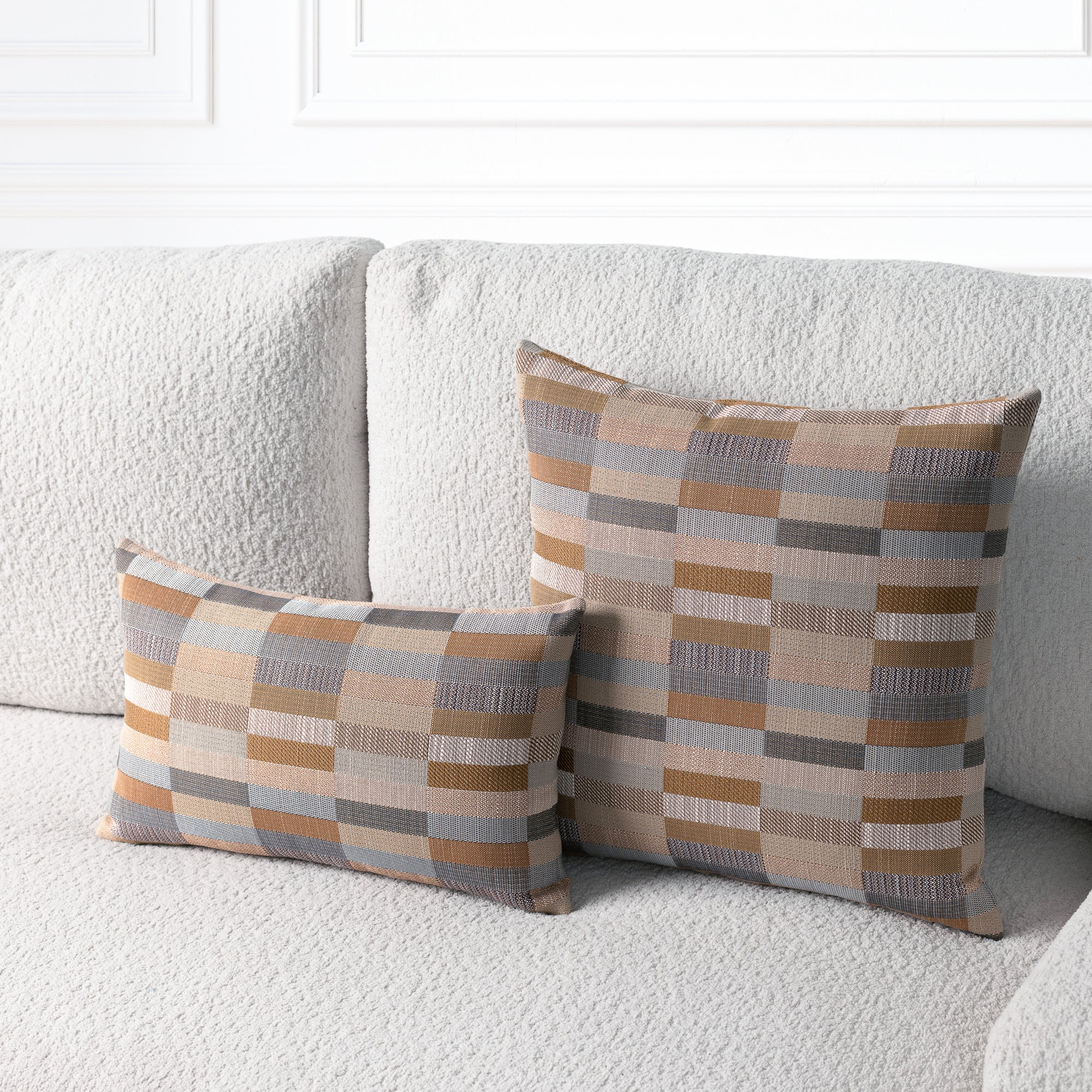 Two rectangular throw pillows with a geometric patchwork plaid pattern in beige, brown, gray, and muted blue, styled on a textured light gray sofa.