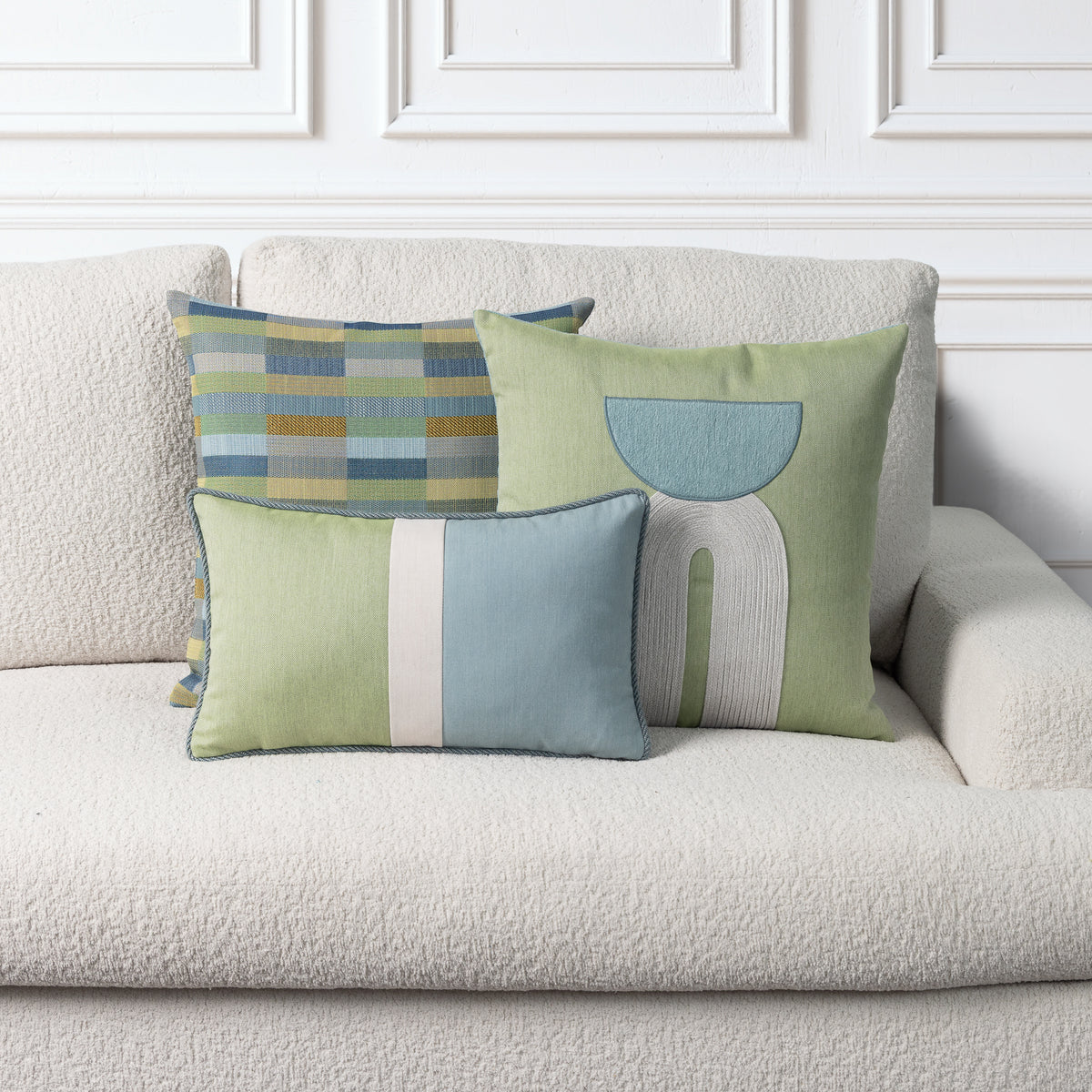Three decorative throw pillows in muted green, light blue, and beige tones arranged on a textured cream sofa against a white paneled wall.
