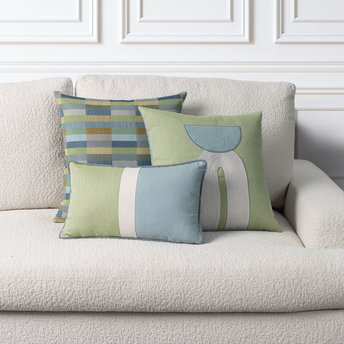 Three decorative throw pillows in muted green, blue, beige, and gray tones arranged on a cream boucle sofa against a white paneled wall.