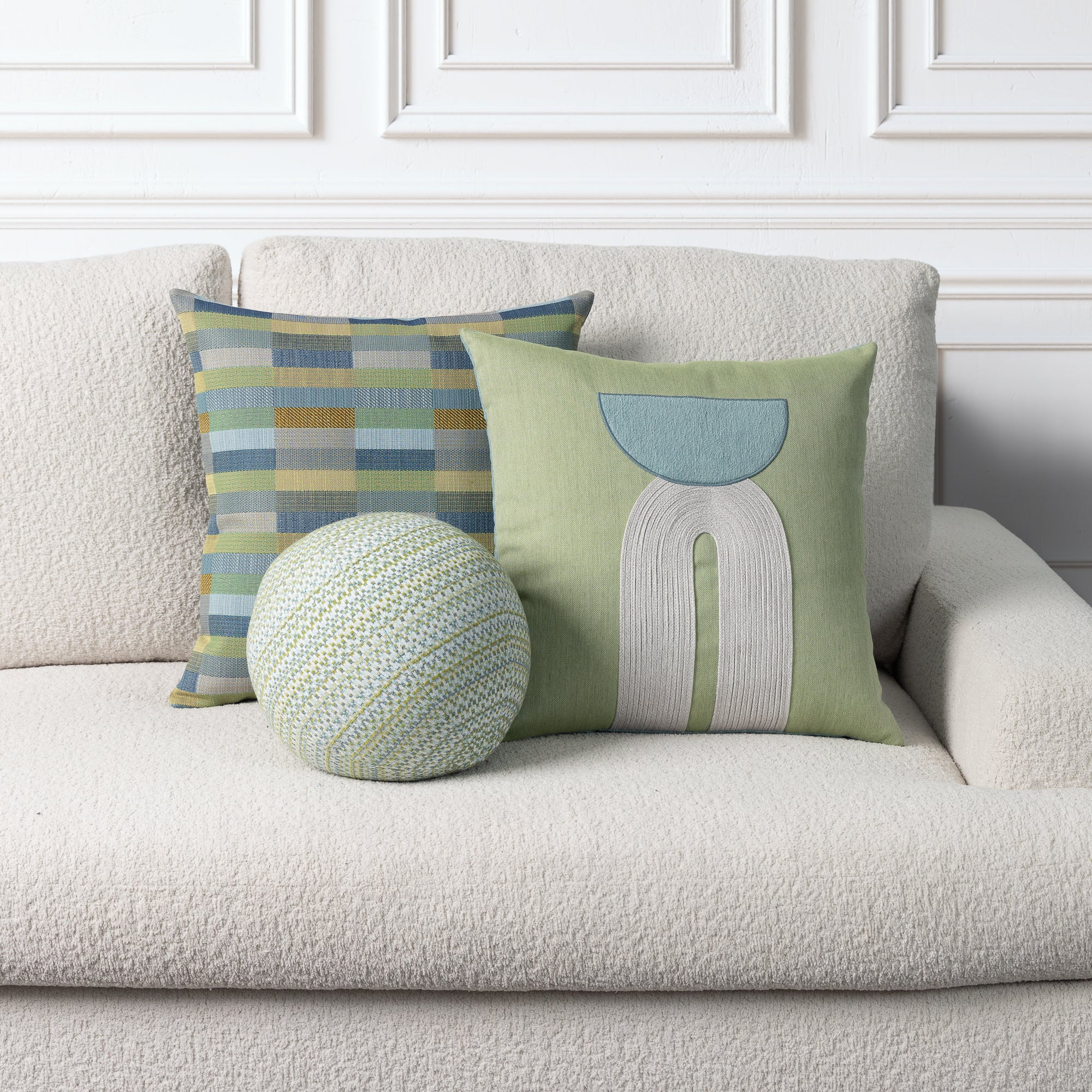 Three decorative throw pillows arranged on a textured cream boucle sofa against a white paneled wall. One square pillow has a green base with a light blue and white abstract arch design, one square pillow has a blue, green, and beige striped pattern, and one round pillow is knitted in light green and cream.
