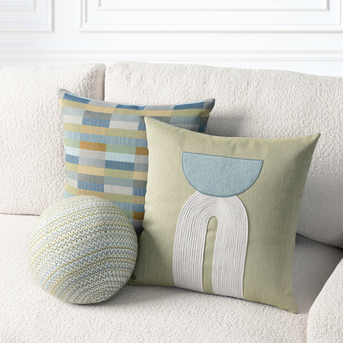 Three decorative throw pillows arranged on a cream boucle sofa: a square pillow with a muted blue, yellow, and beige plaid pattern, a square pillow with an abstract light blue and off-white stitched design on a sage green background, and a round knit pillow in a matching blue, yellow, and beige plaid.