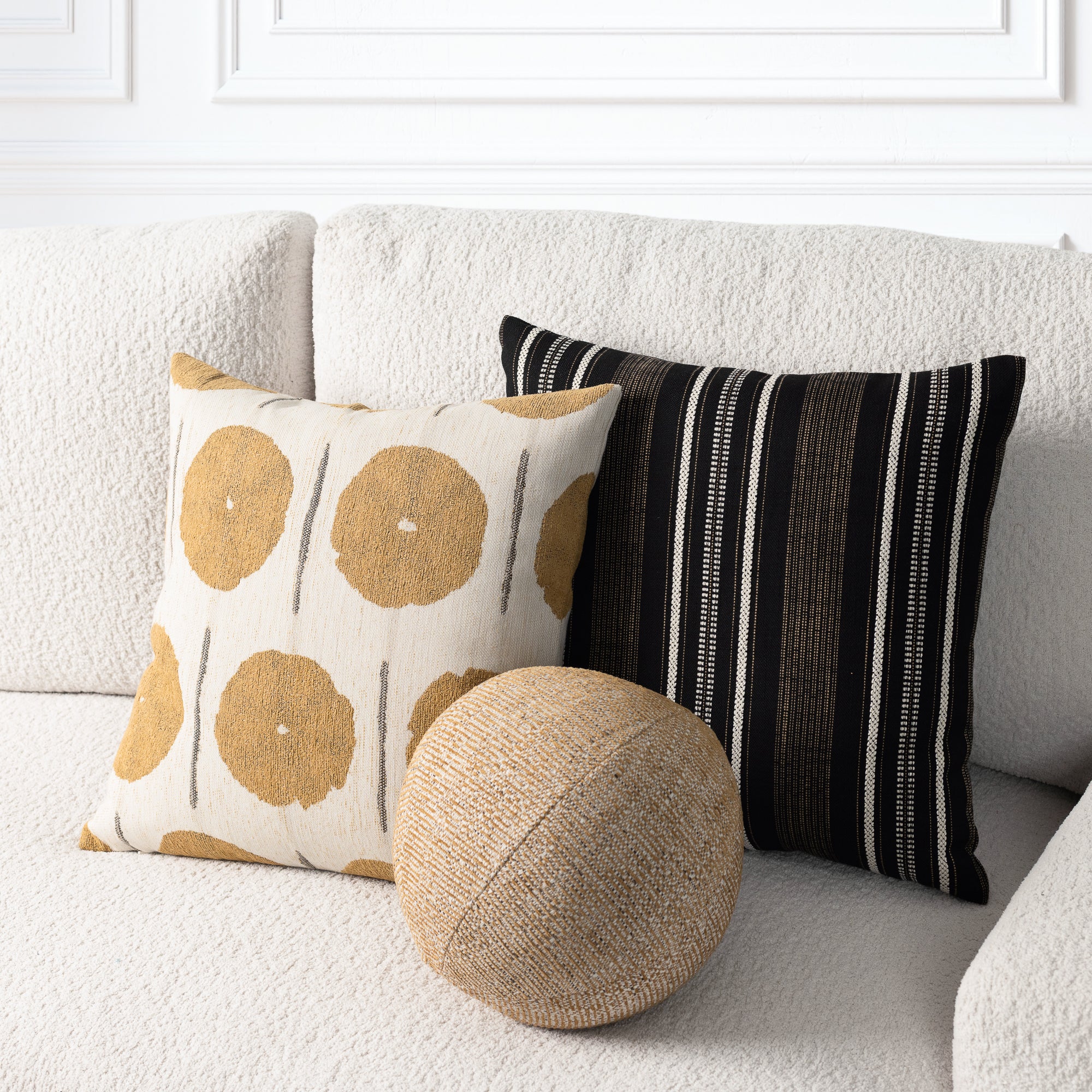 Two decorative throw pillows and a round woven pouf on a textured off-white sofa. One pillow has a cream background with mustard yellow circular motifs and thin gray vertical lines, while the other has black and off-white vertical stripes.