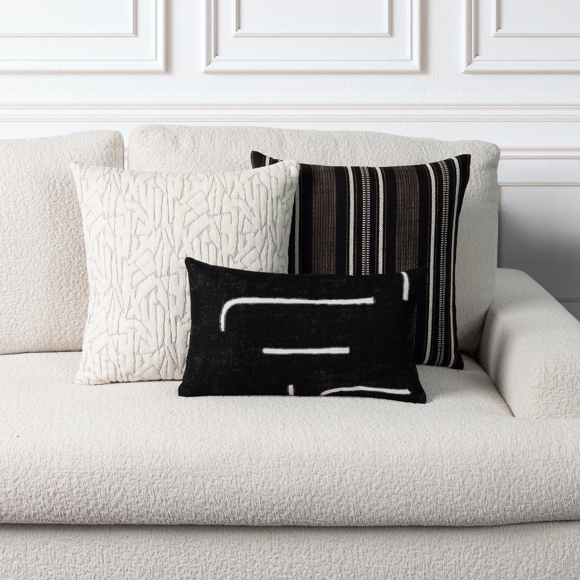 Three decorative throw pillows arranged on a textured off-white sofa against a white paneled wall. The front pillow is black with white abstract lines, the center pillow is cream with a raised abstract pattern, and the back pillow has vertical black, white, and brown stripes.
