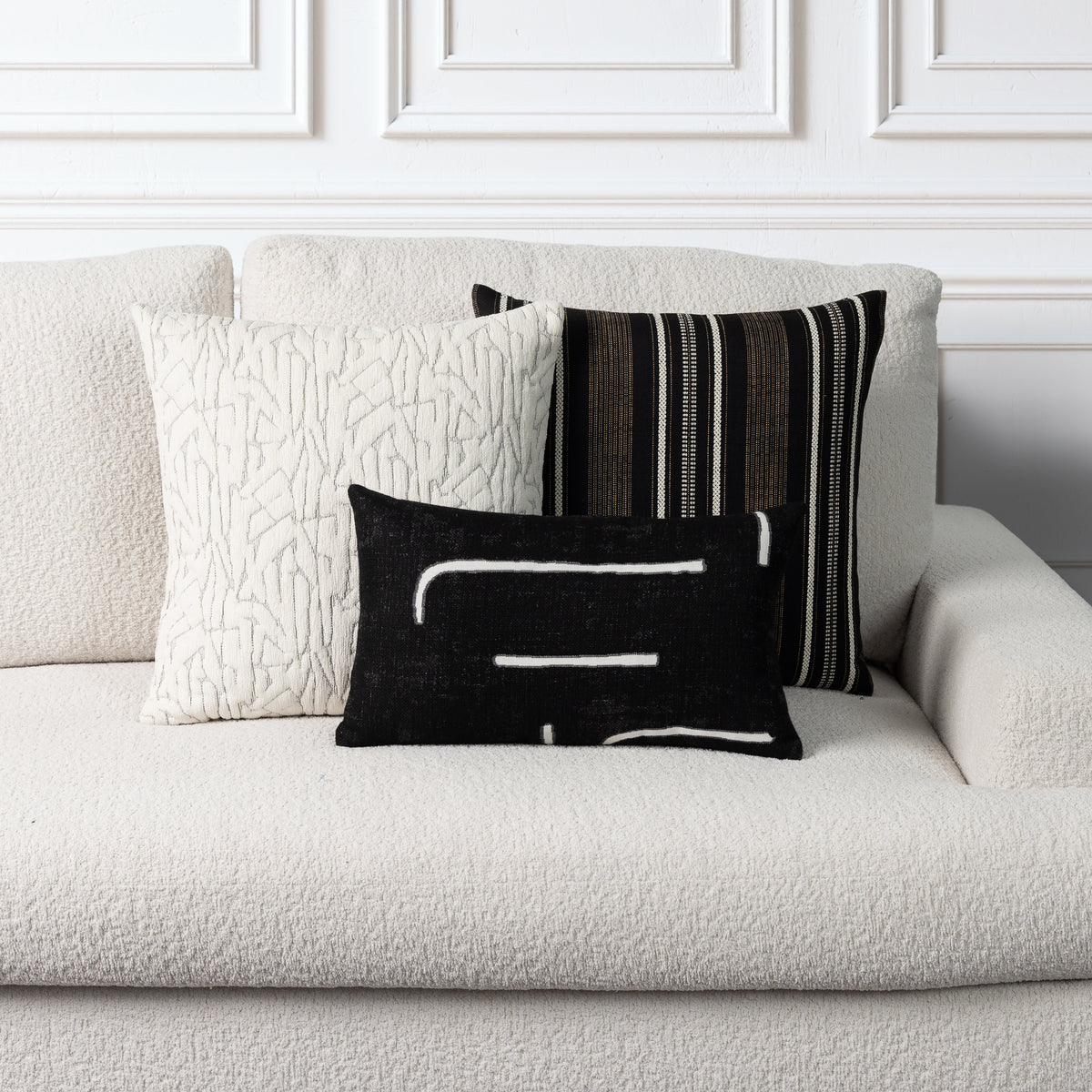 Three decorative throw pillows arranged on a textured off-white sofa against a white paneled wall. The front pillow is black with white abstract lines, the center pillow is cream with a raised abstract pattern, and the back pillow has vertical black, white, and brown stripes.