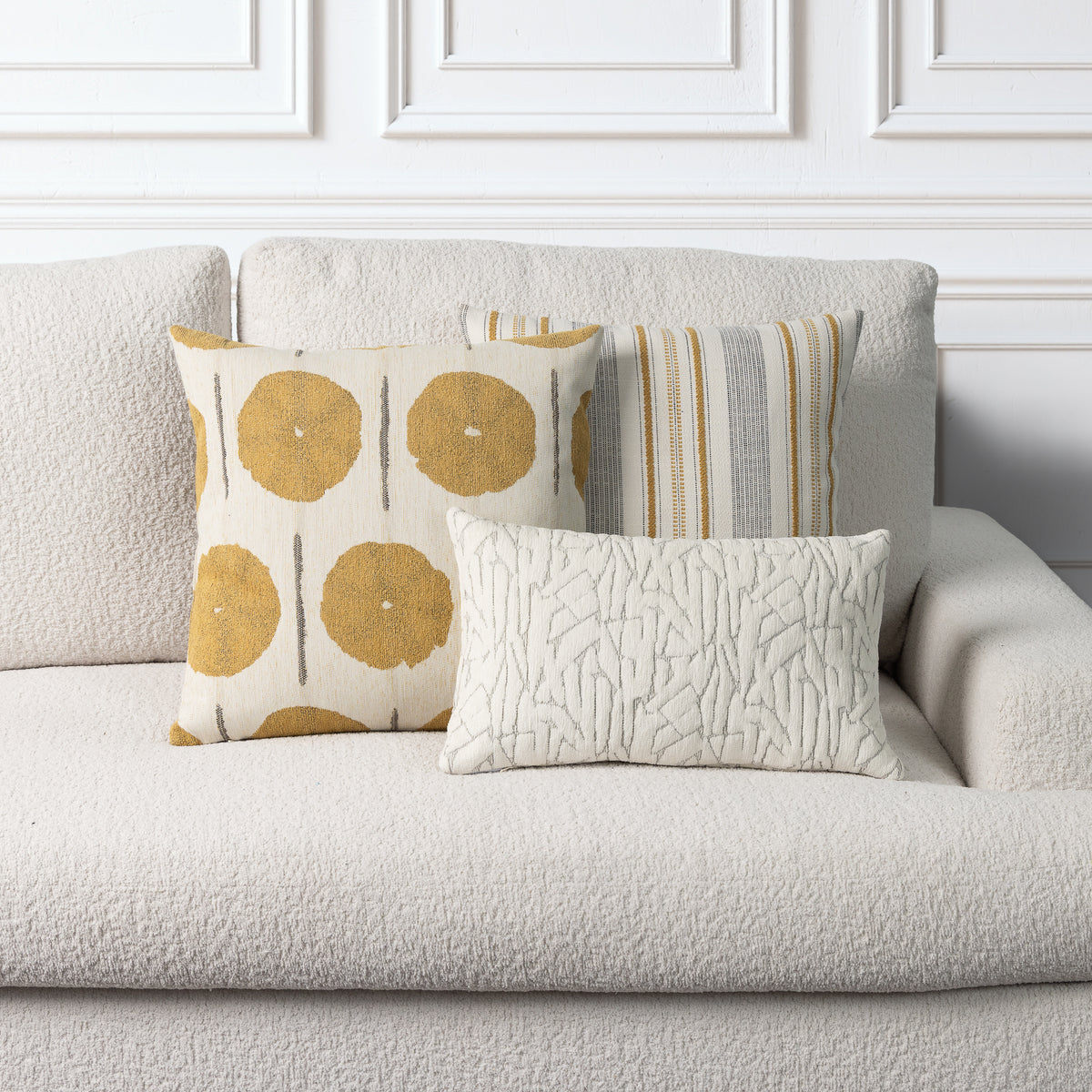 Three decorative throw pillows arranged on a light beige boucle sofa against a white paneled wall. The pillows include a cream pillow with mustard yellow circular motifs, a striped pillow in beige, gray, and yellow, and a cream textured lumbar pillow.