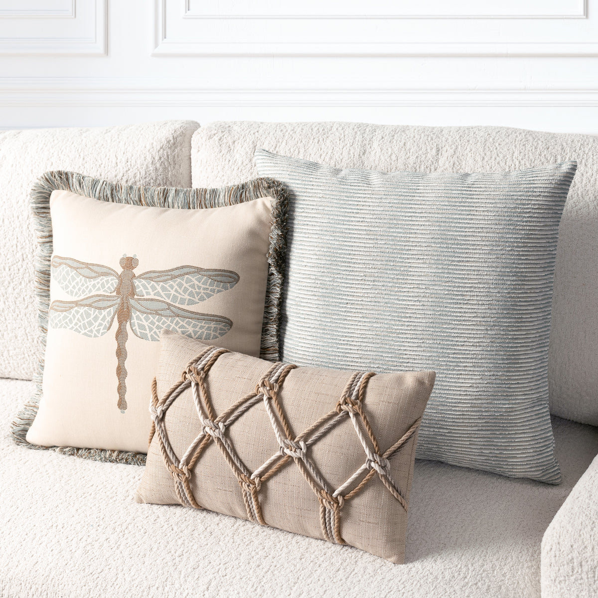 Three decorative throw pillows arranged on a textured white sofa: a large light blue-gray striped pillow, a cream pillow with a dragonfly motif and fringed edge, and a smaller beige pillow with rope lattice detailing.